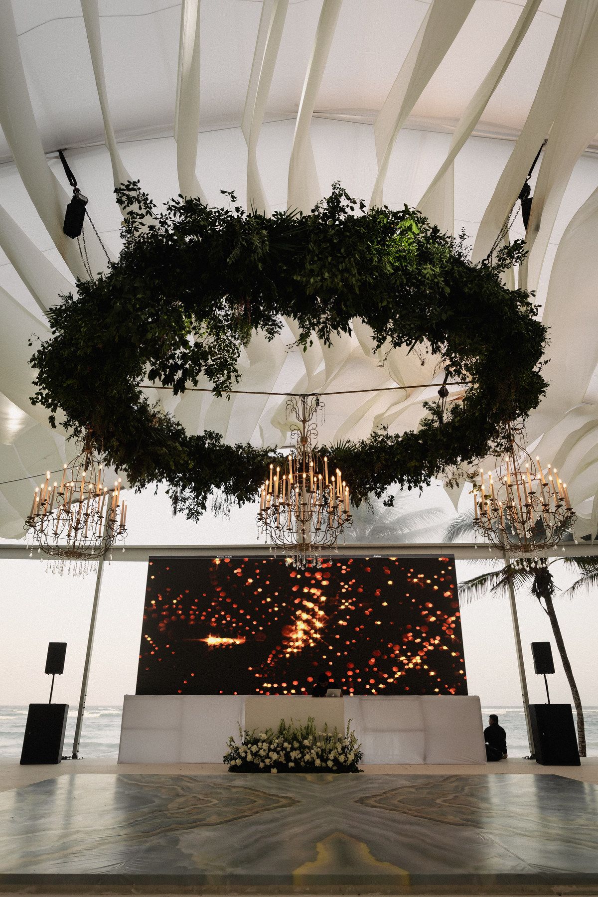 Elegant wedding venue with circular greenery installation and crystal chandeliers over dance floor.