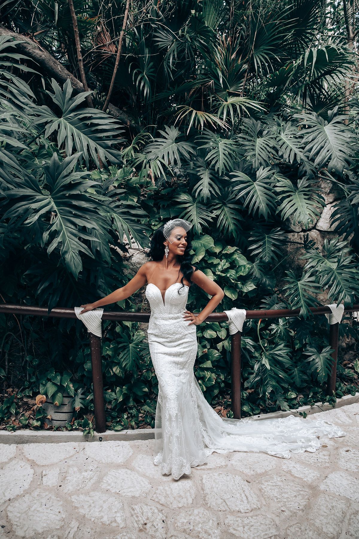 A bride in a fitted lace gown poses against lush tropical foliage.
