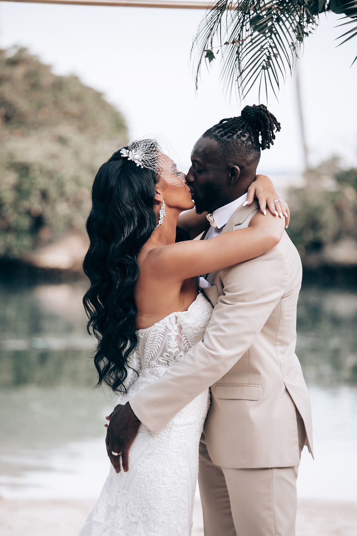 A romantic wedding couple embraces under palm trees beside a tranquil waterfront setting.