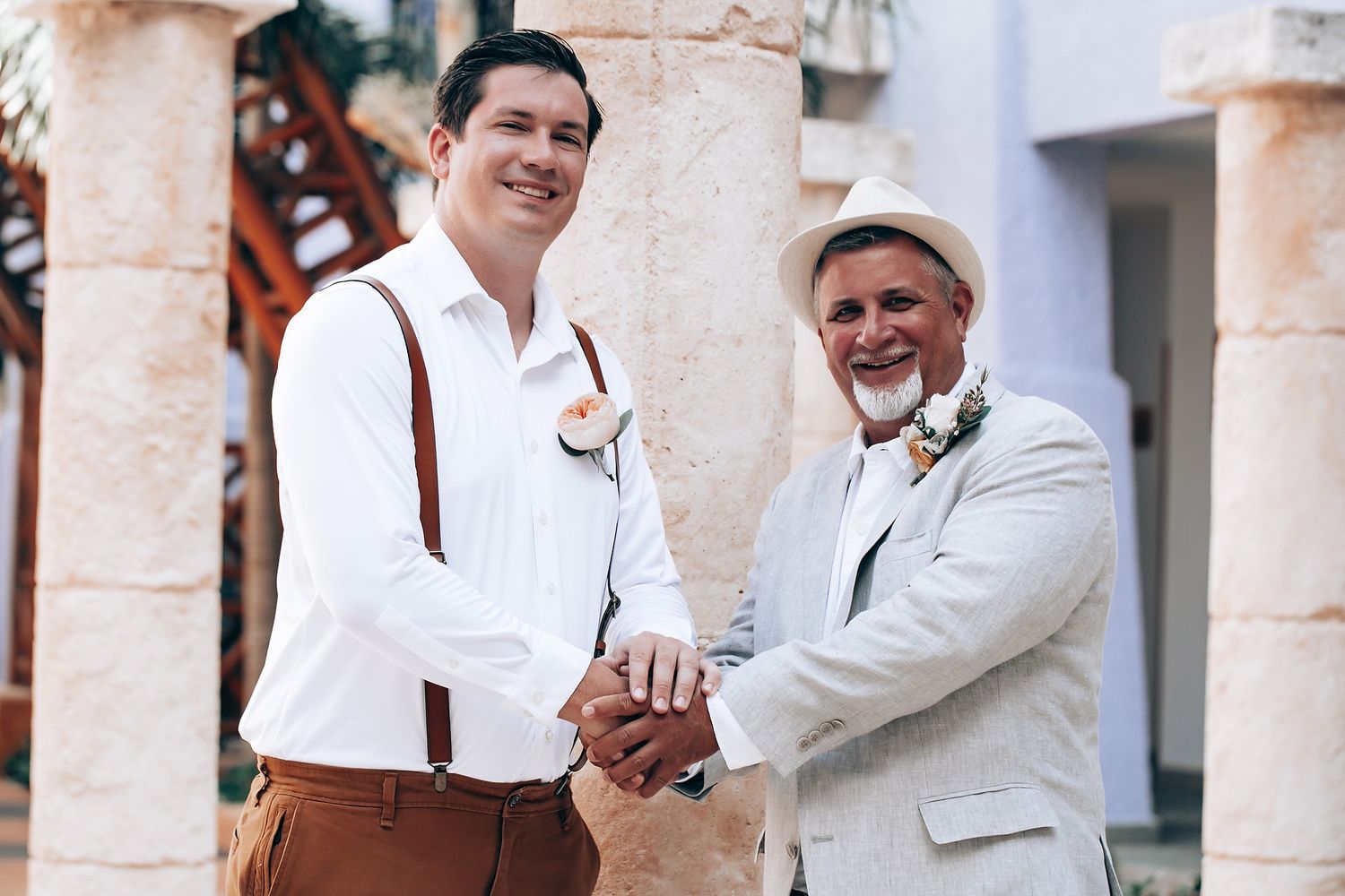 Two men in formal attire share a playful moment in a bright outdoor corridor with stone columns.