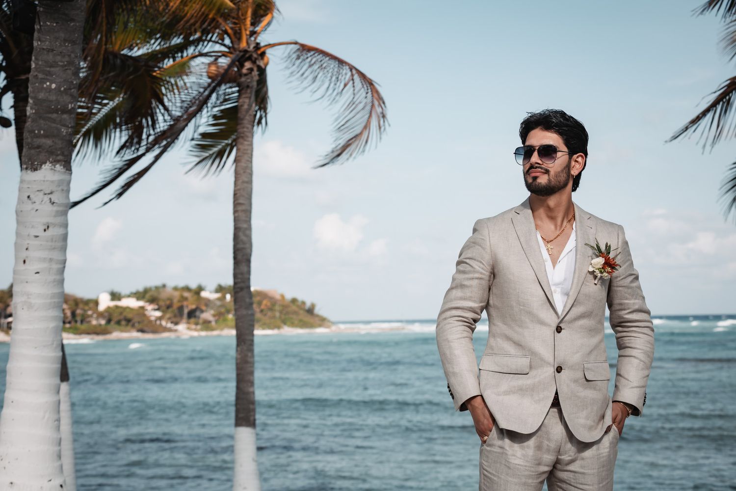 Stylish groom in beige linen suit with sunglasses posing by tropical beach with palm trees.