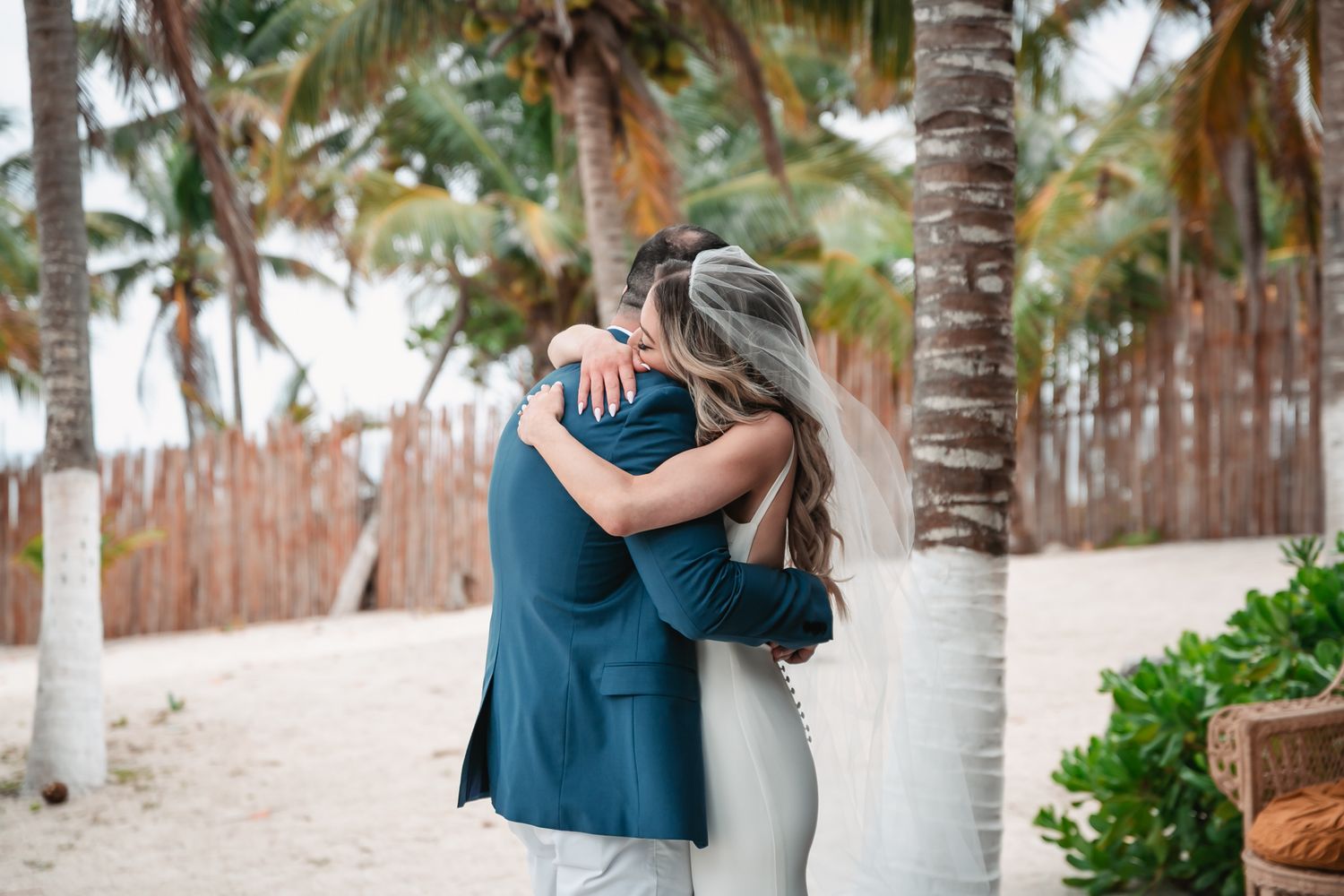 Bride and groom sharing an emotional hug near palm trees on a sandy tropical beach.