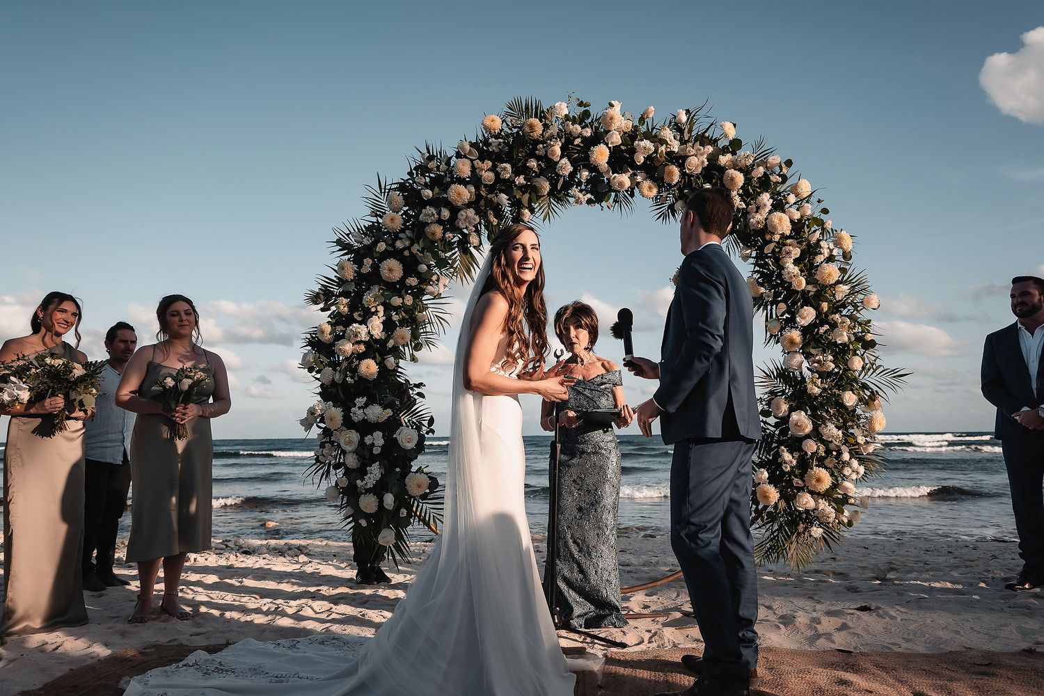 Bride and groom exchange vows under floral arch on beach with ocean backdrop and wedding guests.