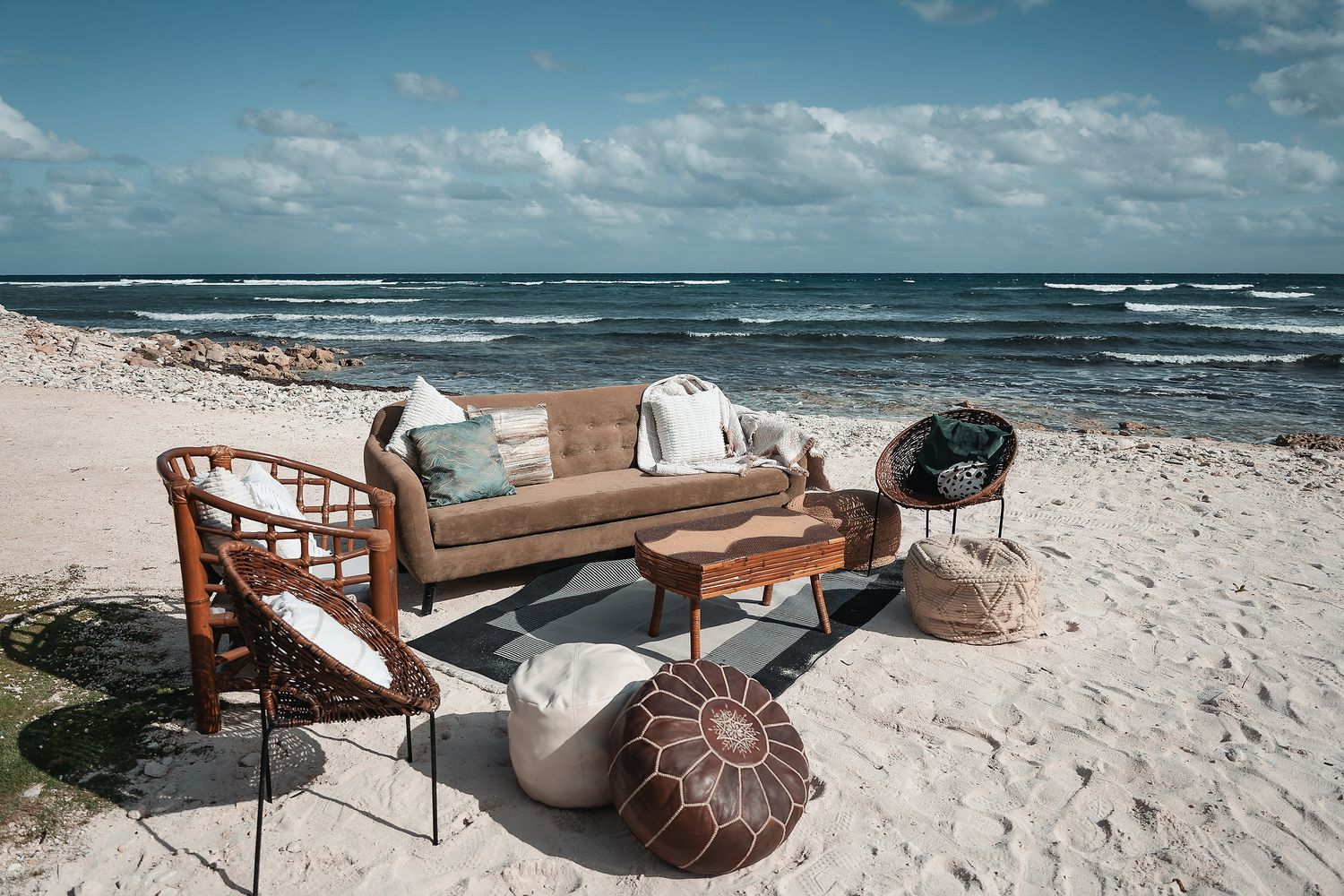 Bohemian beach lounge setup with rattan furniture, cushions, and leather poufs on white sand by the ocean.