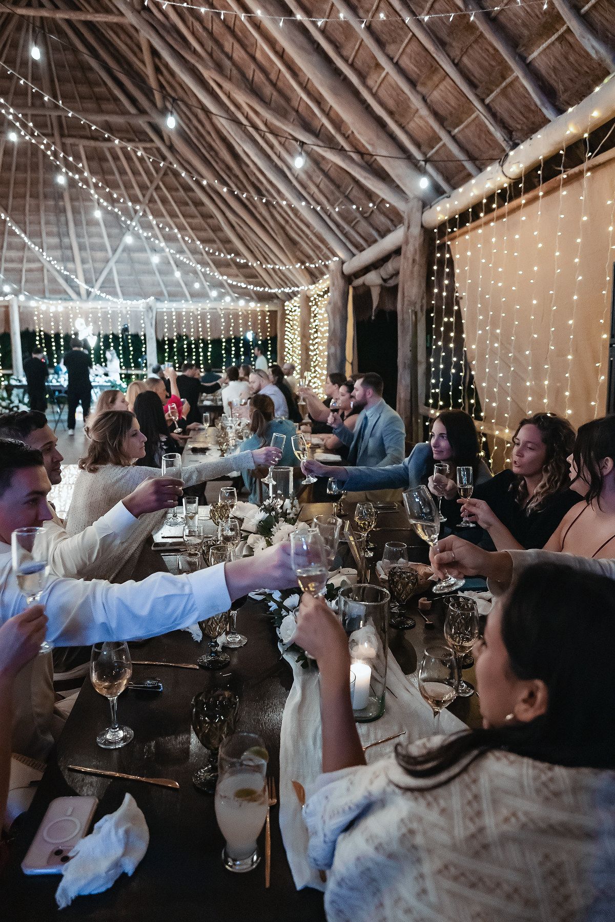 Wedding guests raising glasses for a toast at a rustic barn reception adorned with fairy lights.