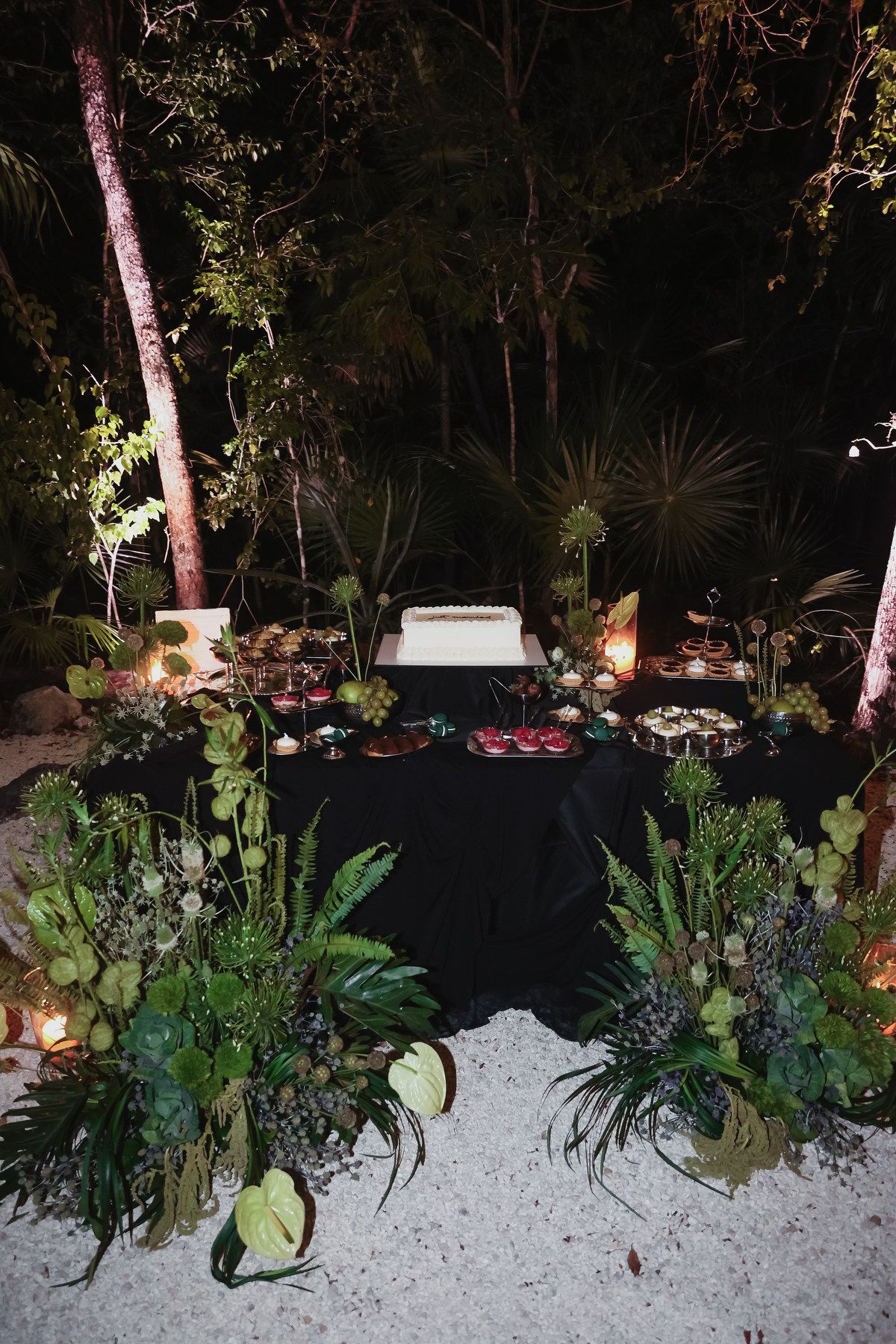 Elegant outdoor dessert table with black linen, tropical greenery, and candlelight in a lush jungle setting at night.