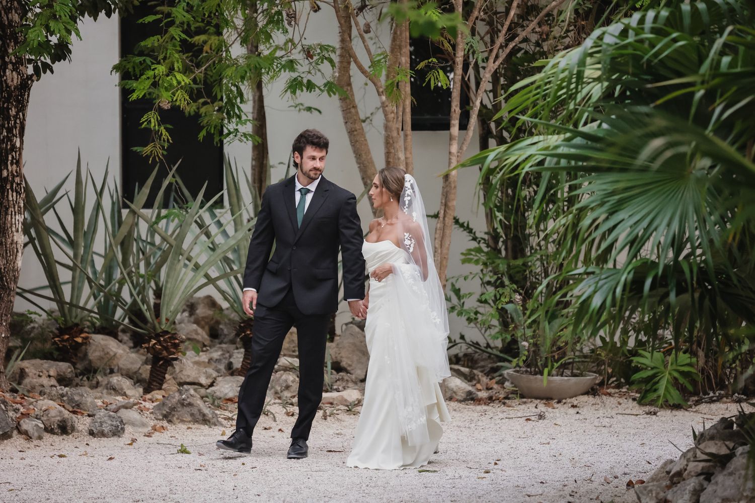 Bride and groom walk hand in hand through tropical garden path on their wedding day.