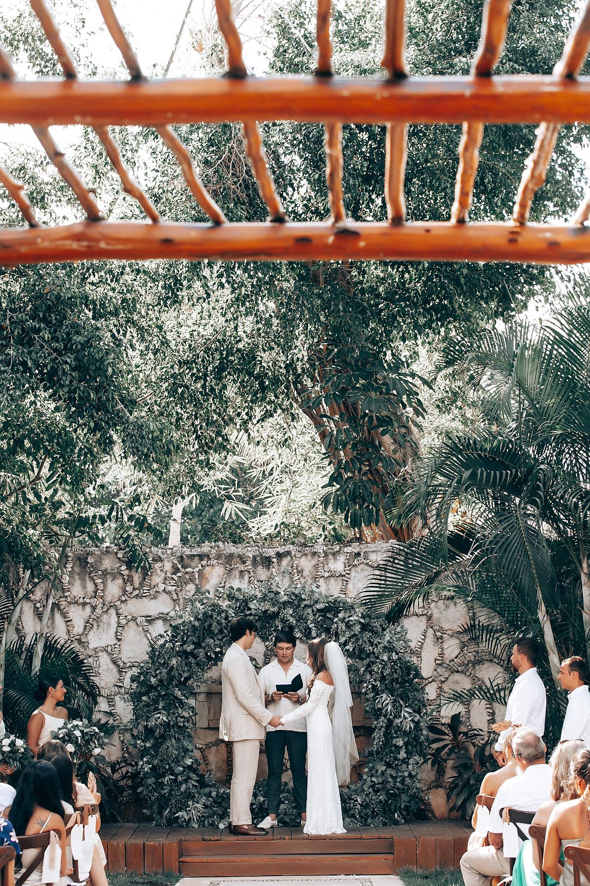 Outdoor wedding ceremony under a wooden pergola with stone wall and tropical garden backdrop.