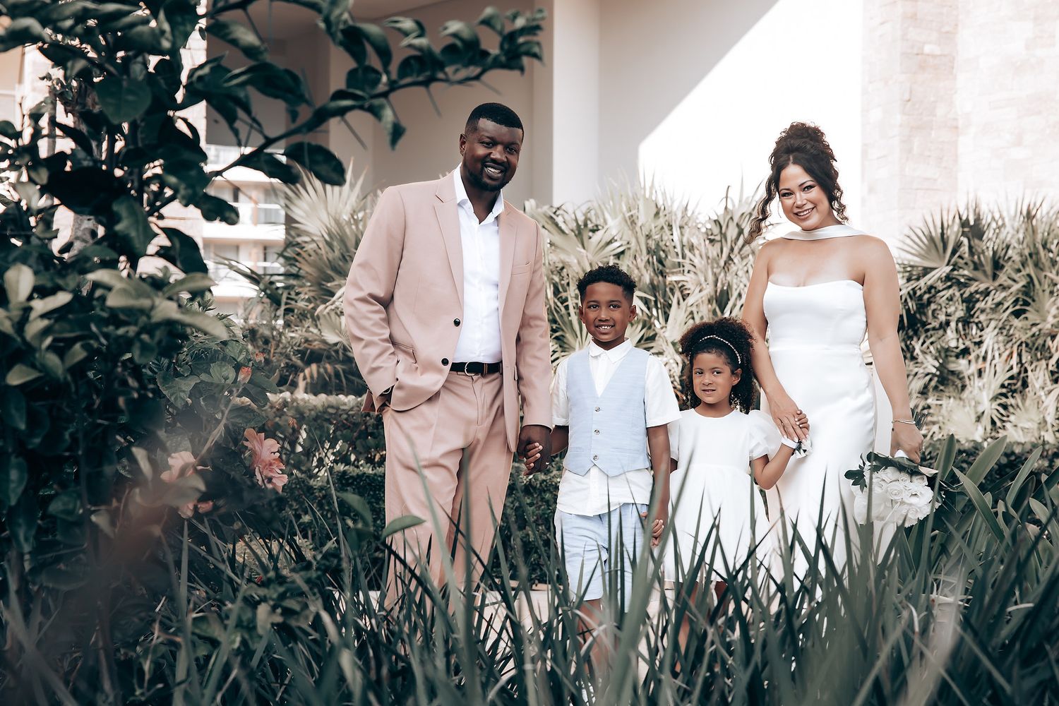 A family poses together in a garden setting wearing coordinated pink and white outfits.