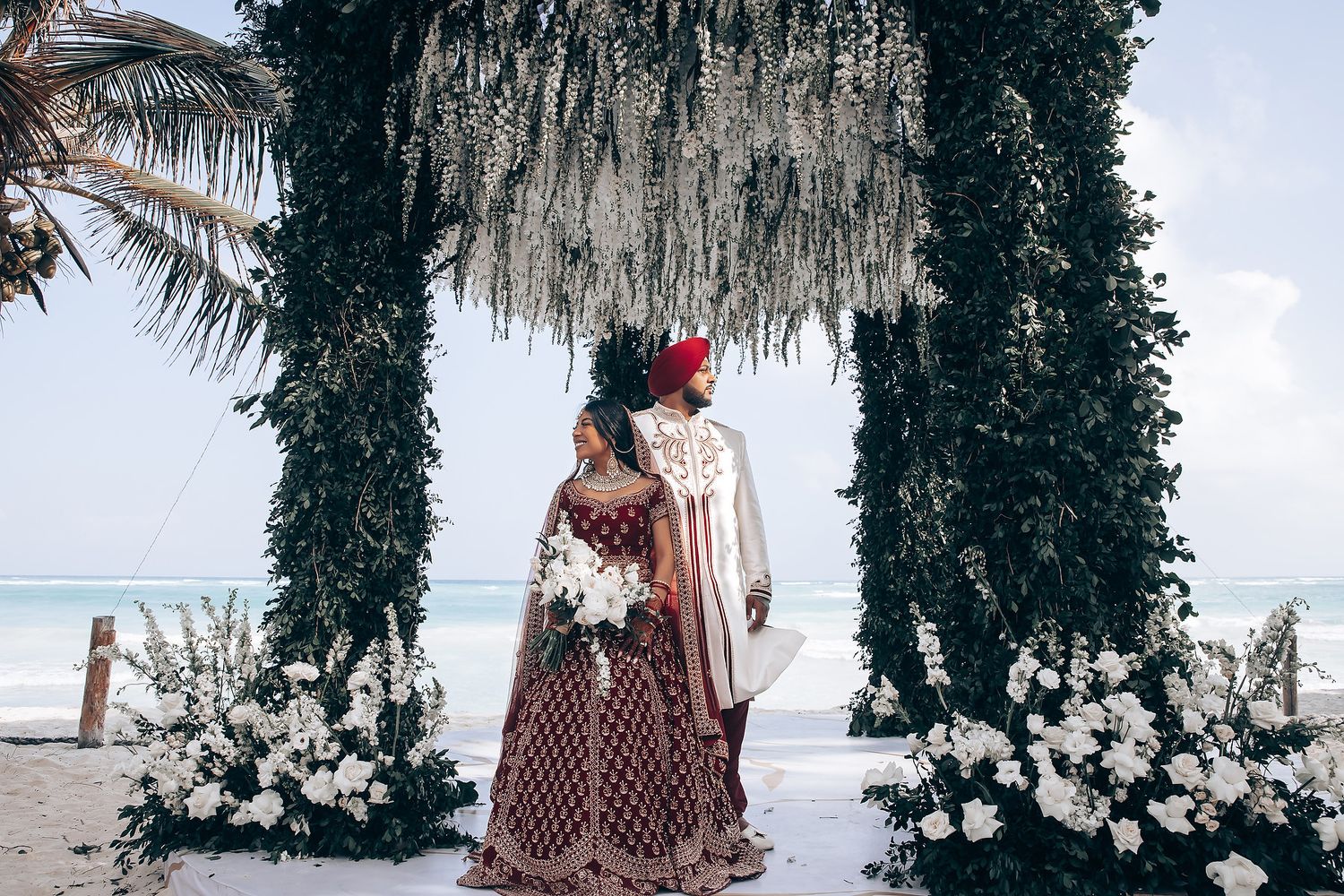 Couple shares romantic moments under cascading white flowers at beach wedding.