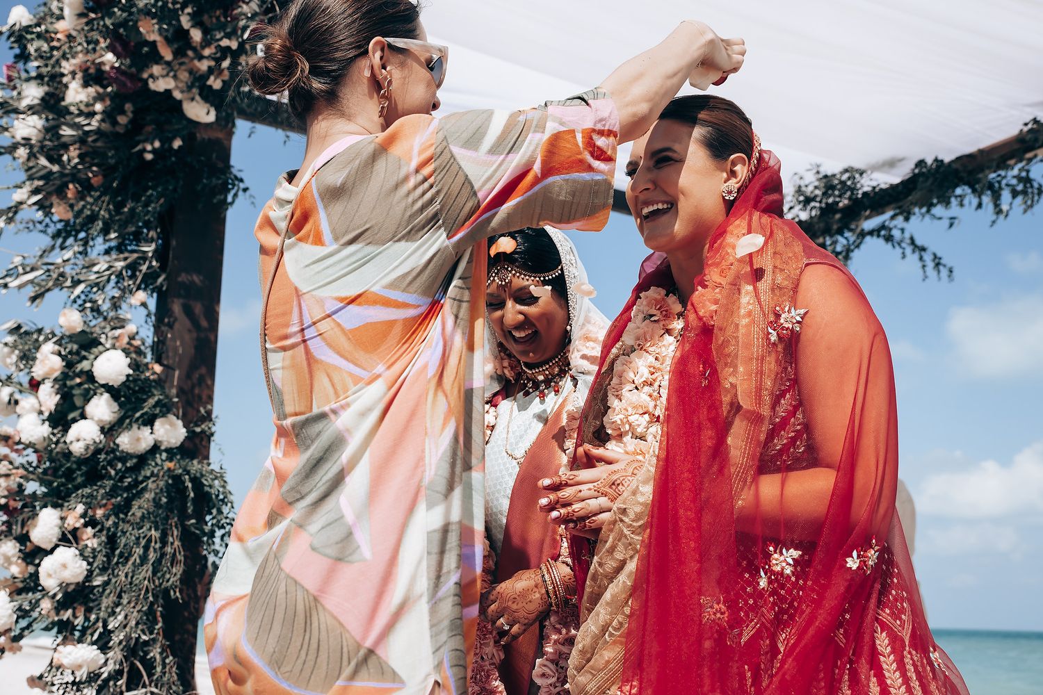 Wedding ceremony participants in traditional Indian attire under floral-decorated canopy.