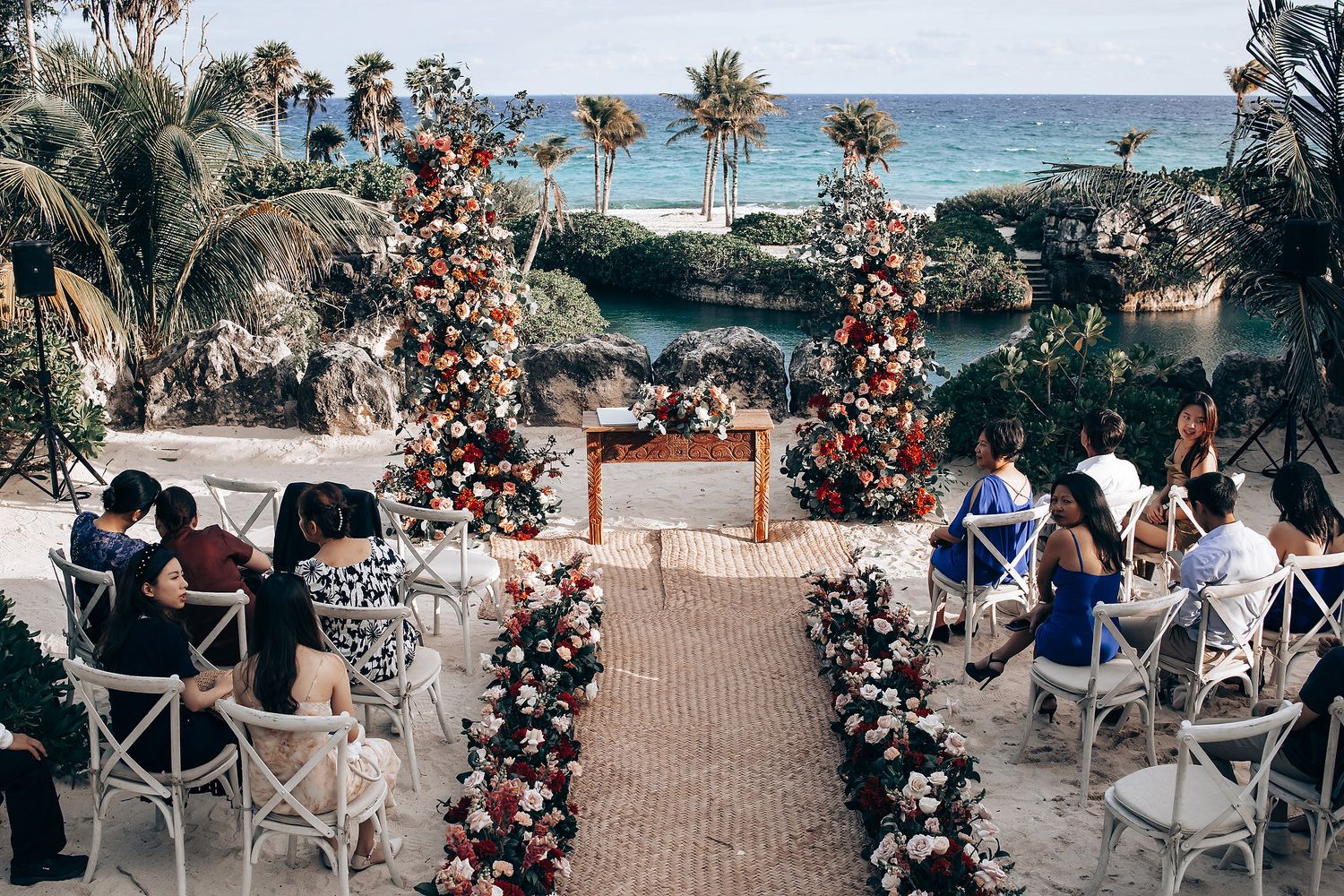 Outdoor beach wedding ceremony setup with floral arch and palm trees against turquoise ocean backdrop.
