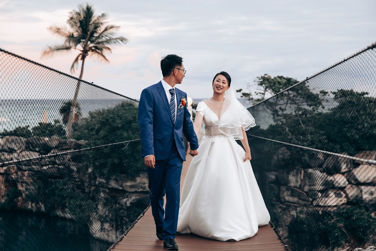 Couple in wedding attire walking on bridge with tropical scenery in background.