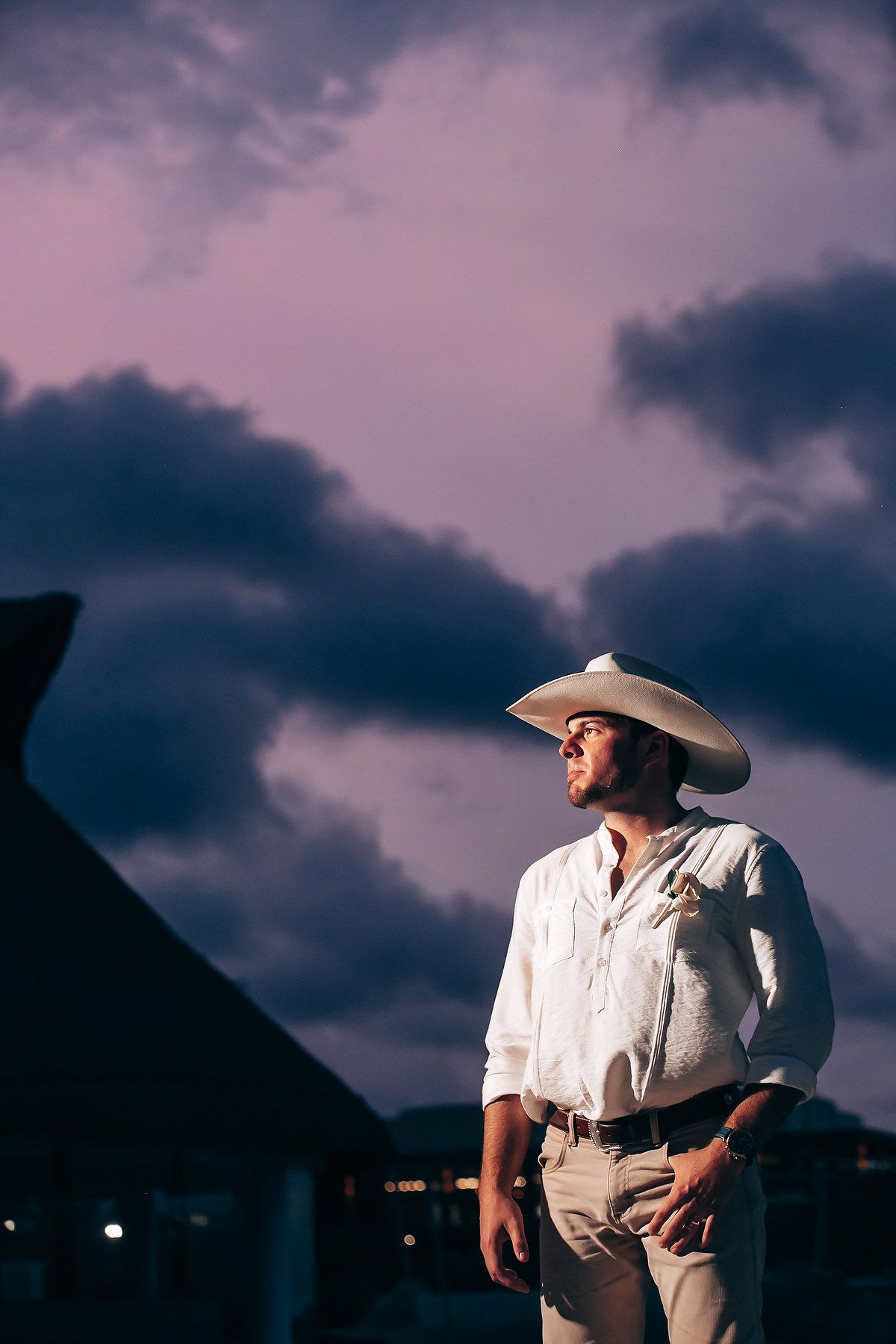 A figure in a white shirt and cowboy hat stands against a dramatic purple sunset sky at dusk.