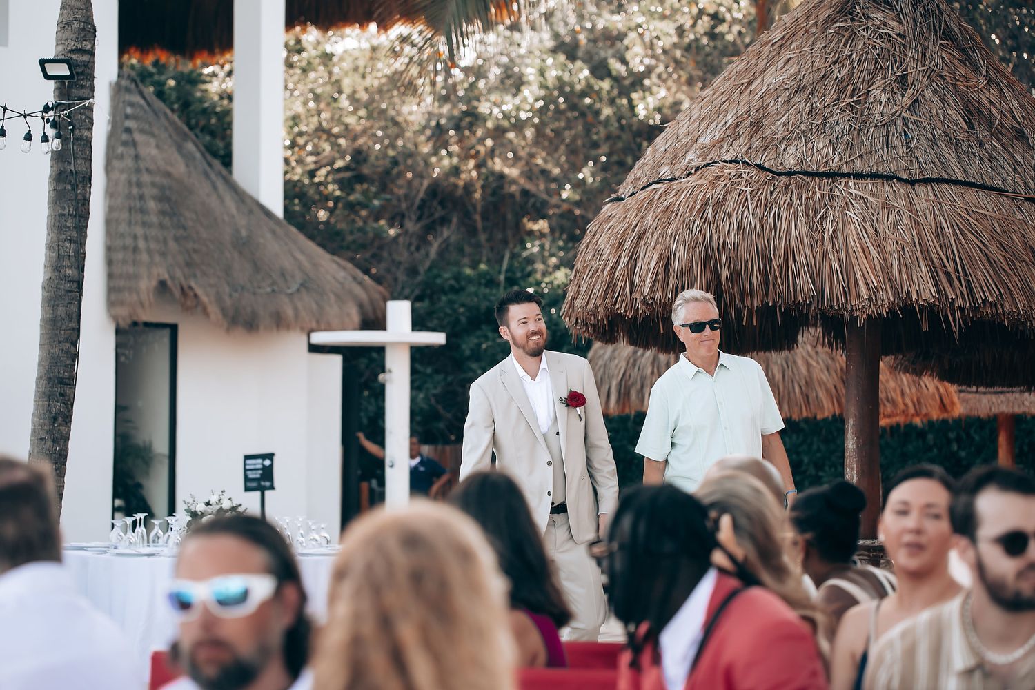 Wedding guests seated in white chairs on a tropical beach with palm trees and thatched umbrellas.