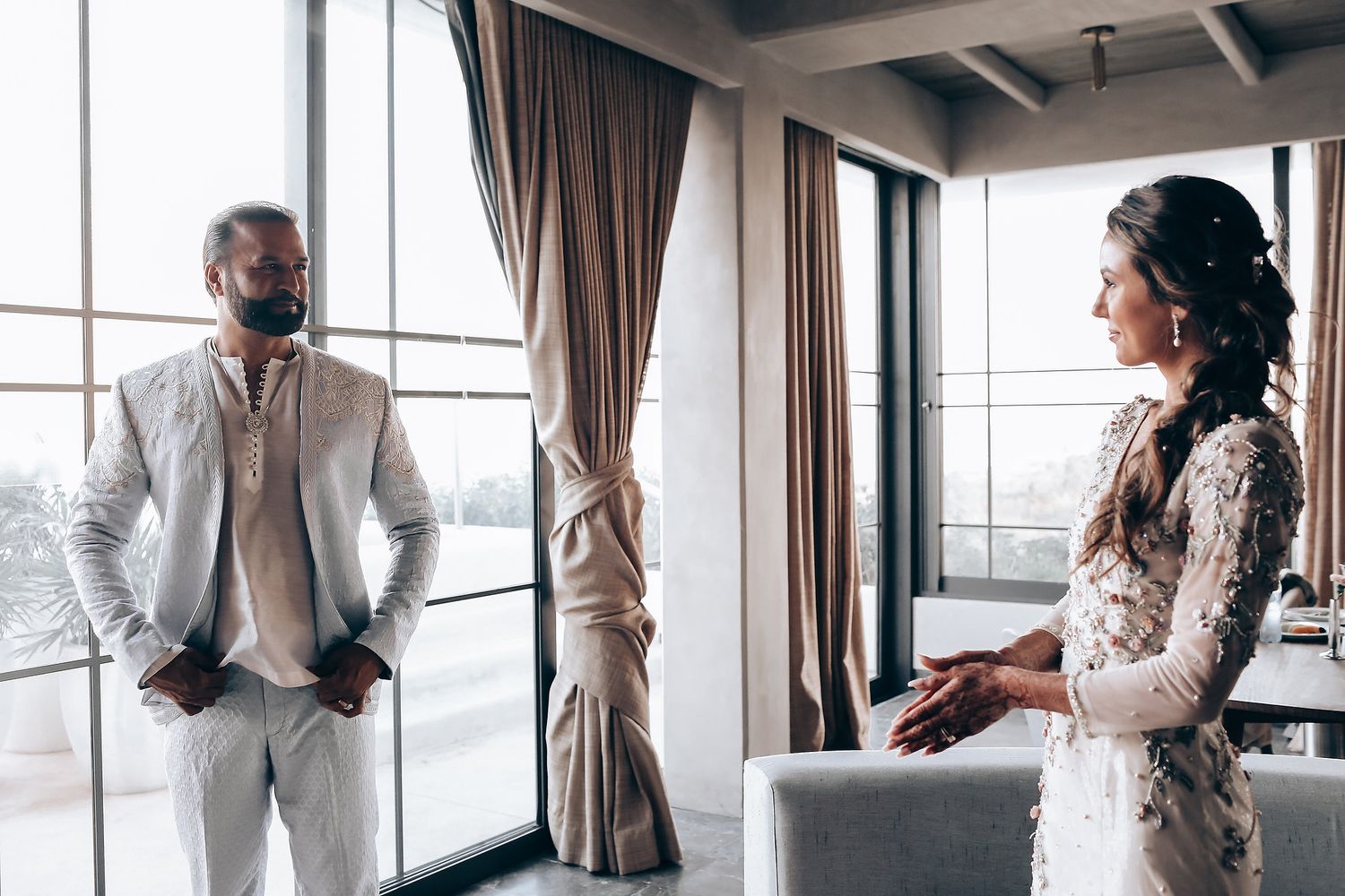 A wedding couple shares intimate moments in a modern hotel room with large windows and elegant curtains.