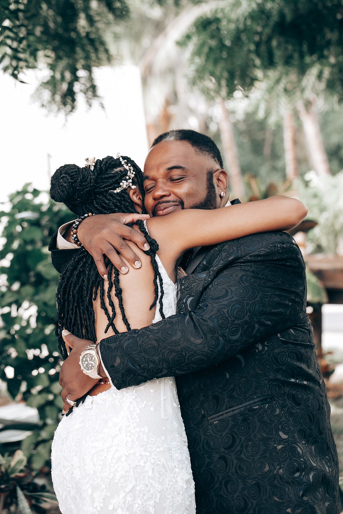A couple shares an intimate embrace outdoors during their wedding celebration, with trees and greenery in the background.