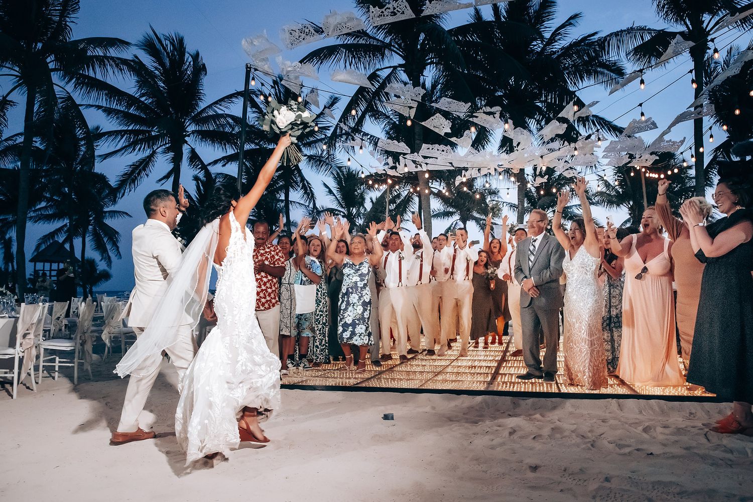 Wedding guests in coordinated attire celebrate on a scenic beach with palm trees at sunset.