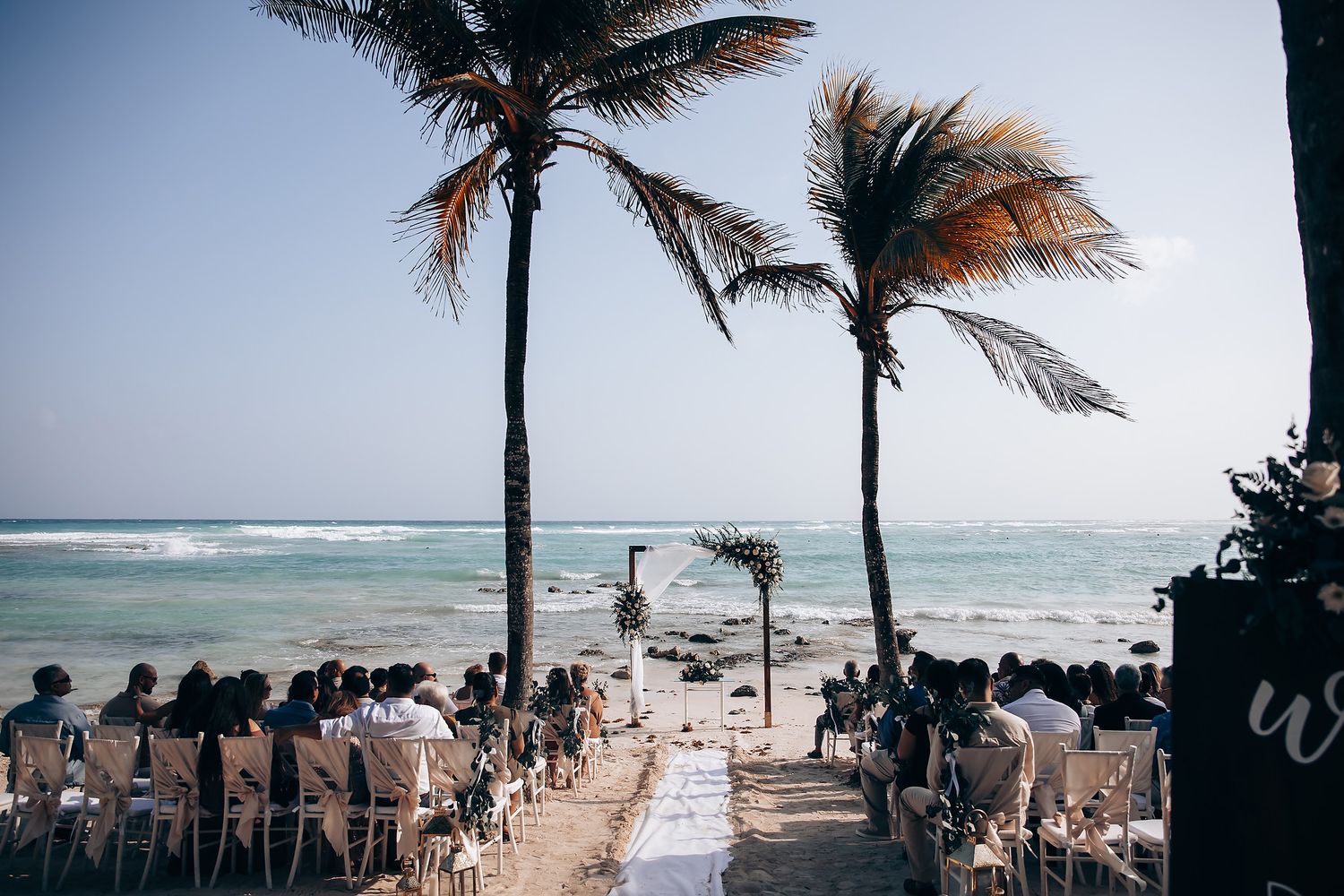Palm trees sway in the wind along a tropical beach with white sand and turquoise waters under cloudy skies.