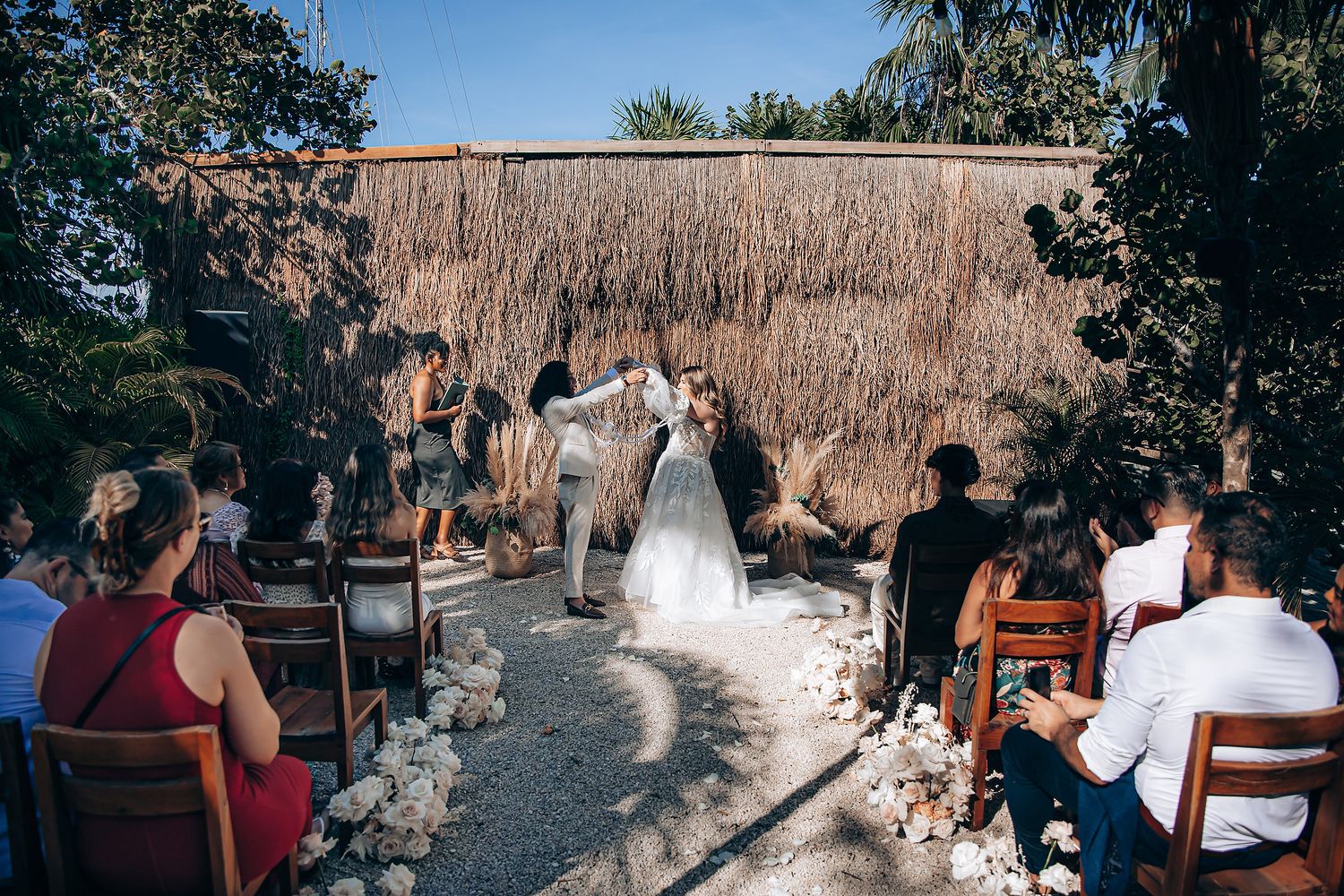 Wedding guests gather at an outdoor reception venue with thatched roof structures and stone pathways.
