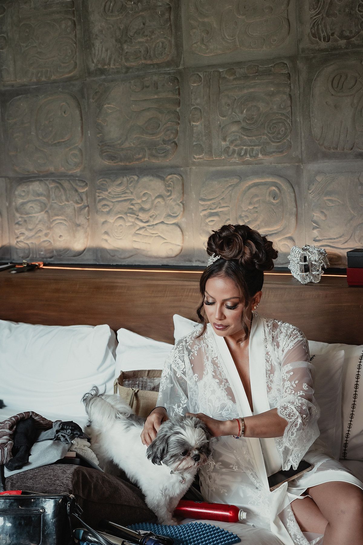 A bride in a white lace gown relaxes on a couch with a fluffy white dog during a wedding day photoshoot.