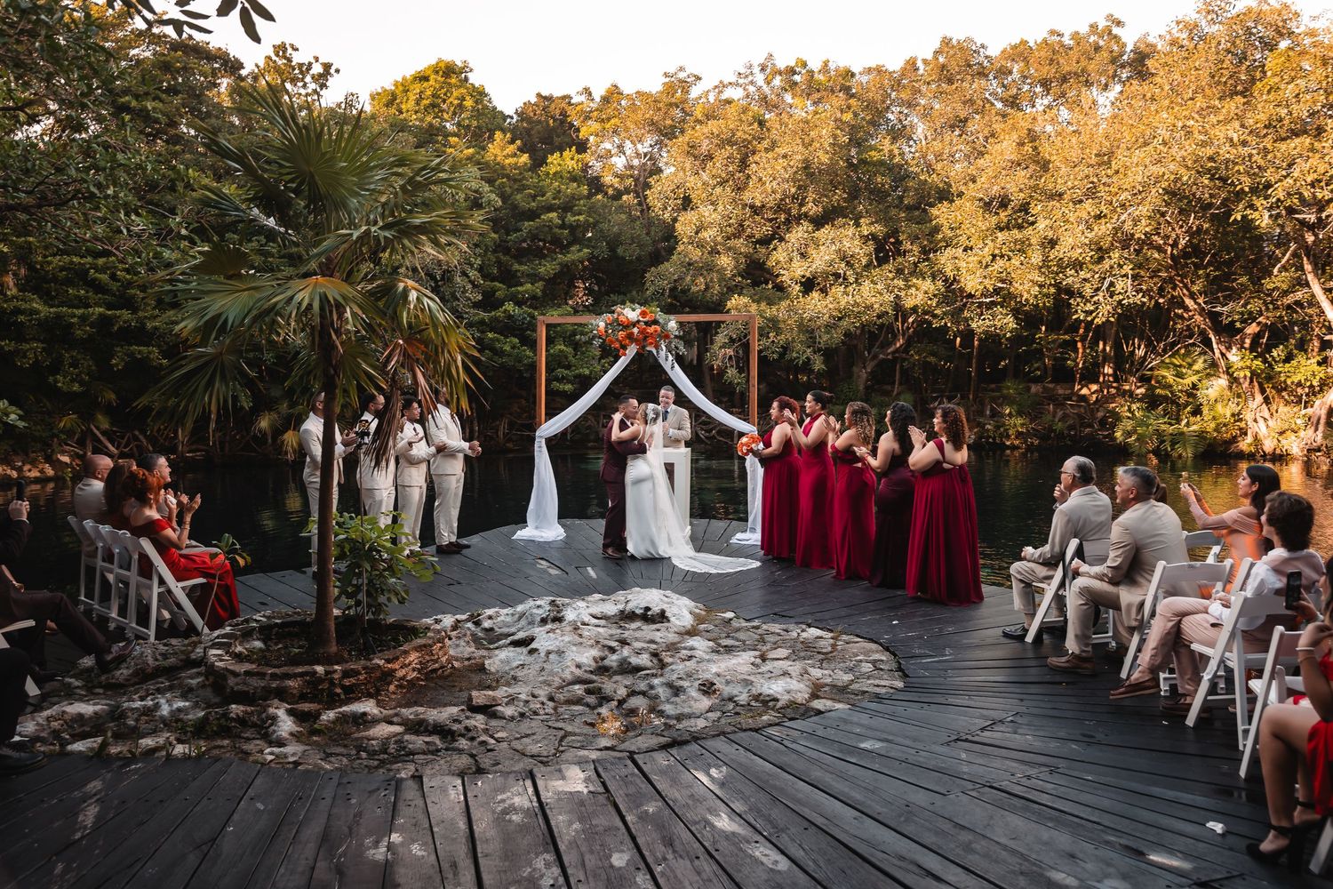 Wedding party poses together on rustic wooden deck surrounded by autumn foliage at outdoor venue.