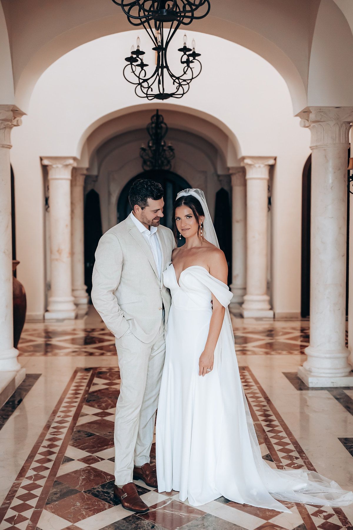 Couple shares romantic moment under elegant arches with wrought iron chandeliers in marble hallway.