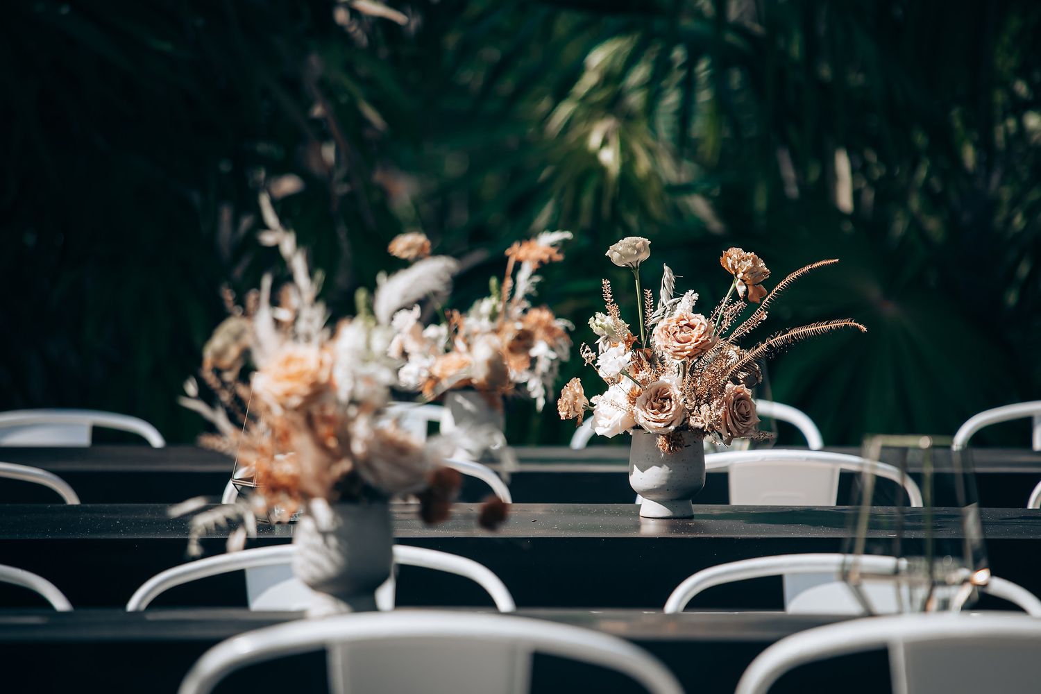 A row of elegant table settings with blush pink floral centerpieces and white chairs at an outdoor wedding reception.