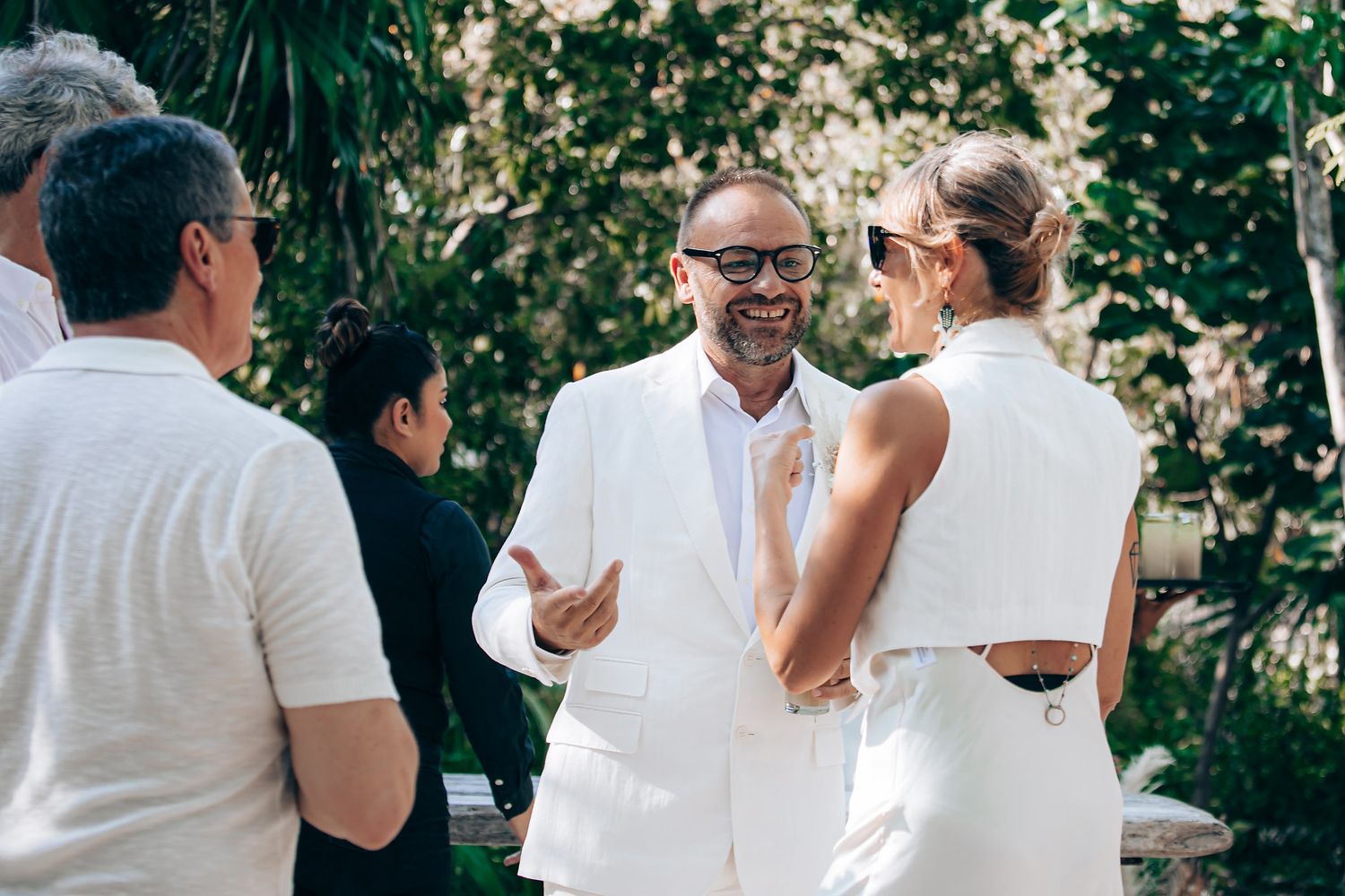 Wedding guests in white attire mingle and socialize during an outdoor garden reception celebration.