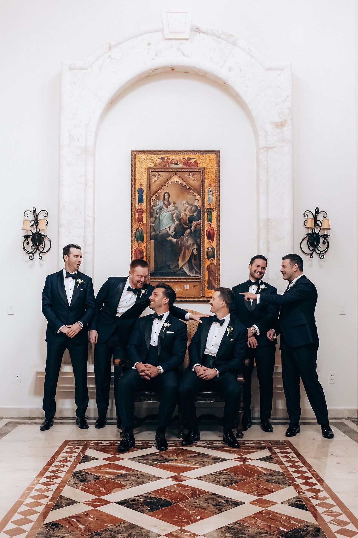 Groomsmen in black tuxedos pose together beneath an ornate religious painting and arched white wall with decorative sconces.