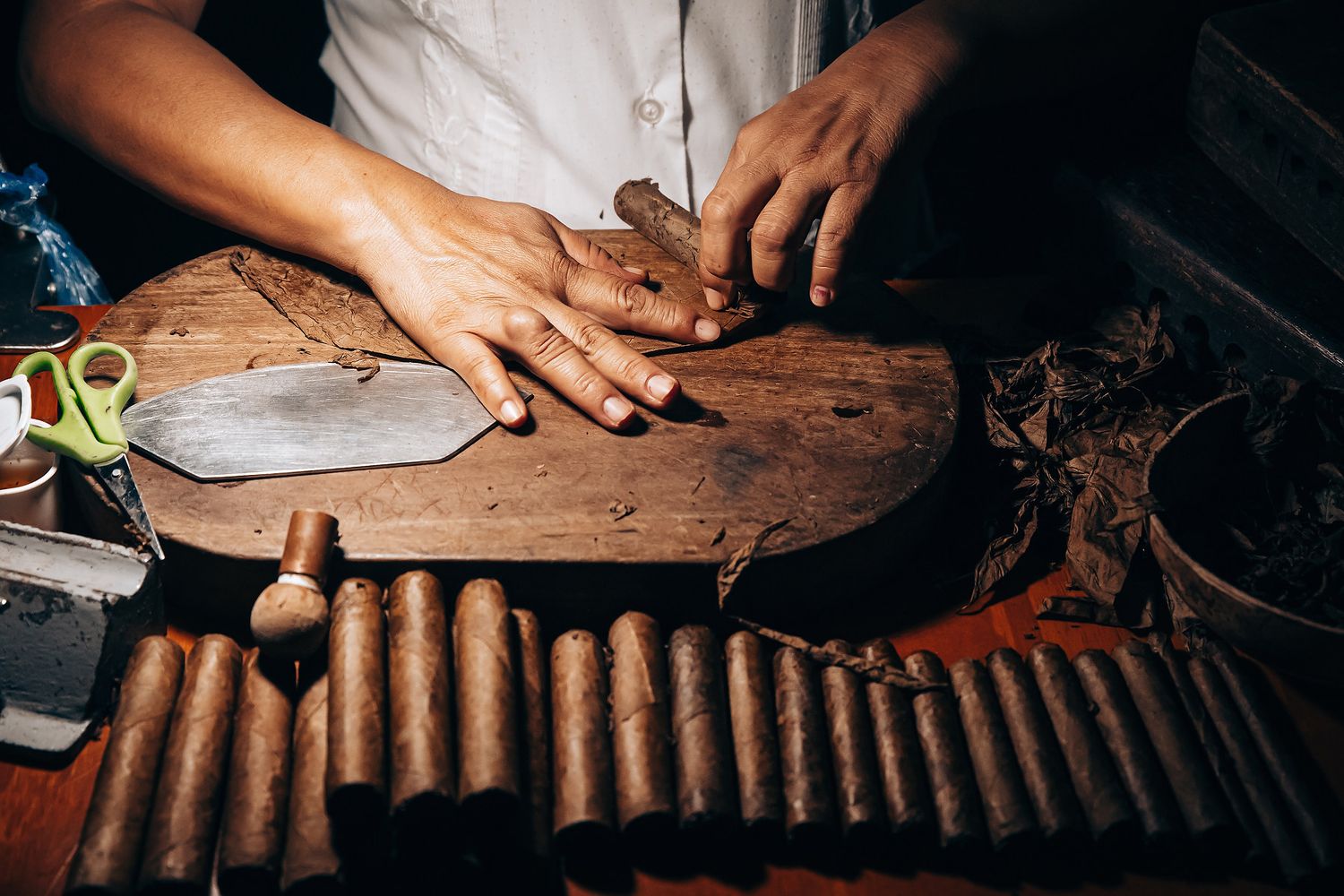 Hands rolling traditional cigars on wooden table with tobacco leaves and tools in dark lighting conditions.