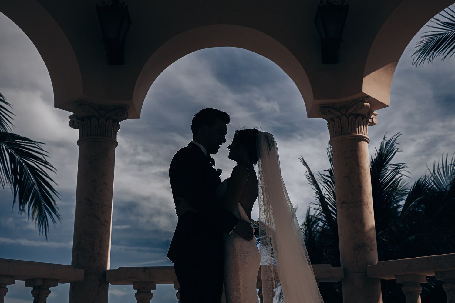 Silhouetted couple embraces under arched columns with palm trees and dramatic cloudy sky in the background.