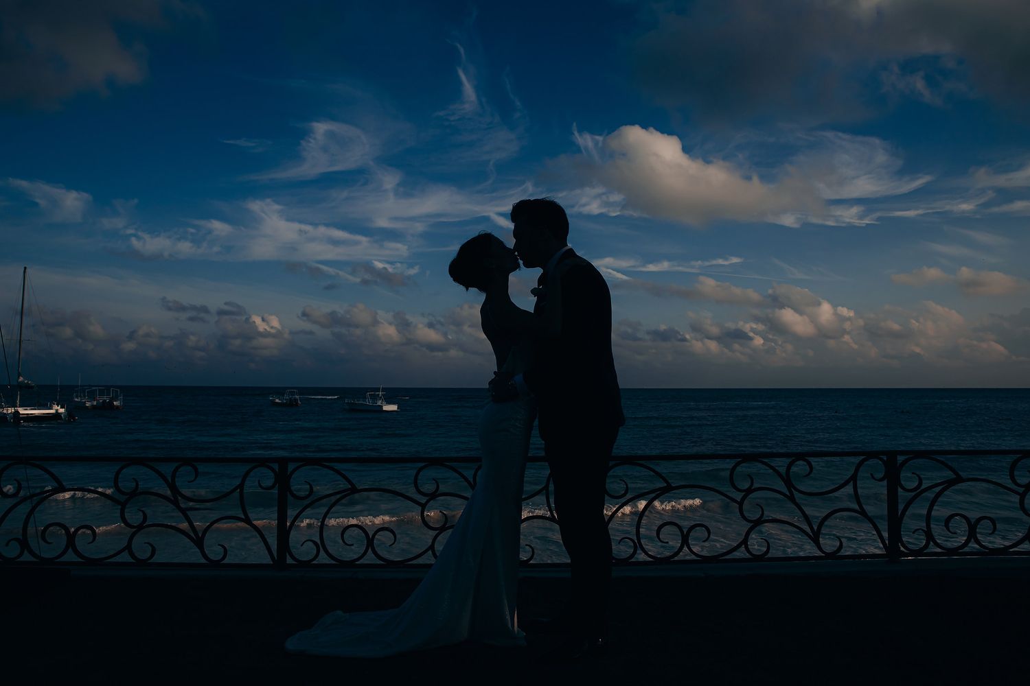 Silhouette of couple embracing on balcony overlooking ocean at sunset with boats in distance.