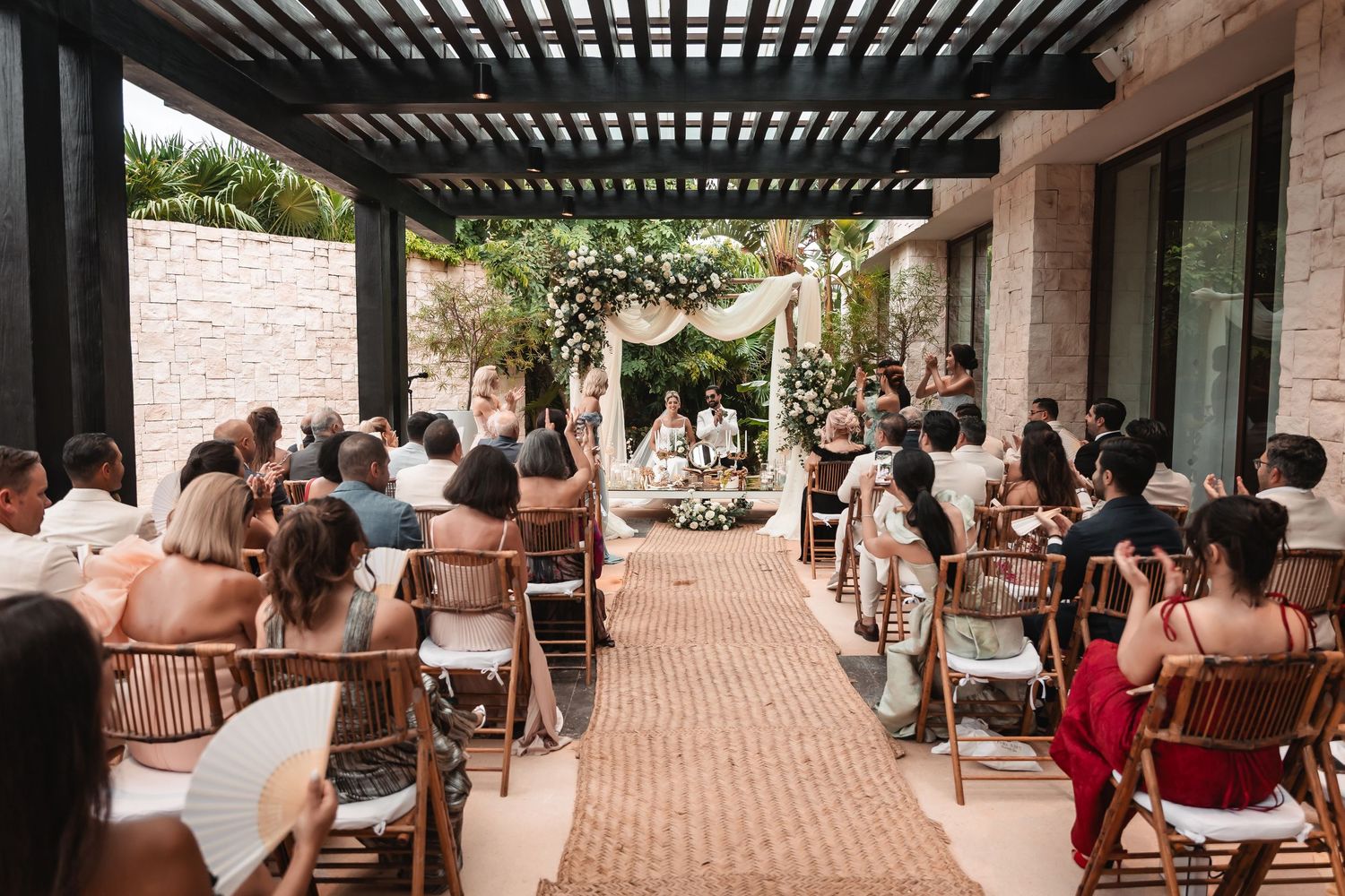 Panoramic view of wedding guests seated at an outdoor reception under a pergola-covered patio.