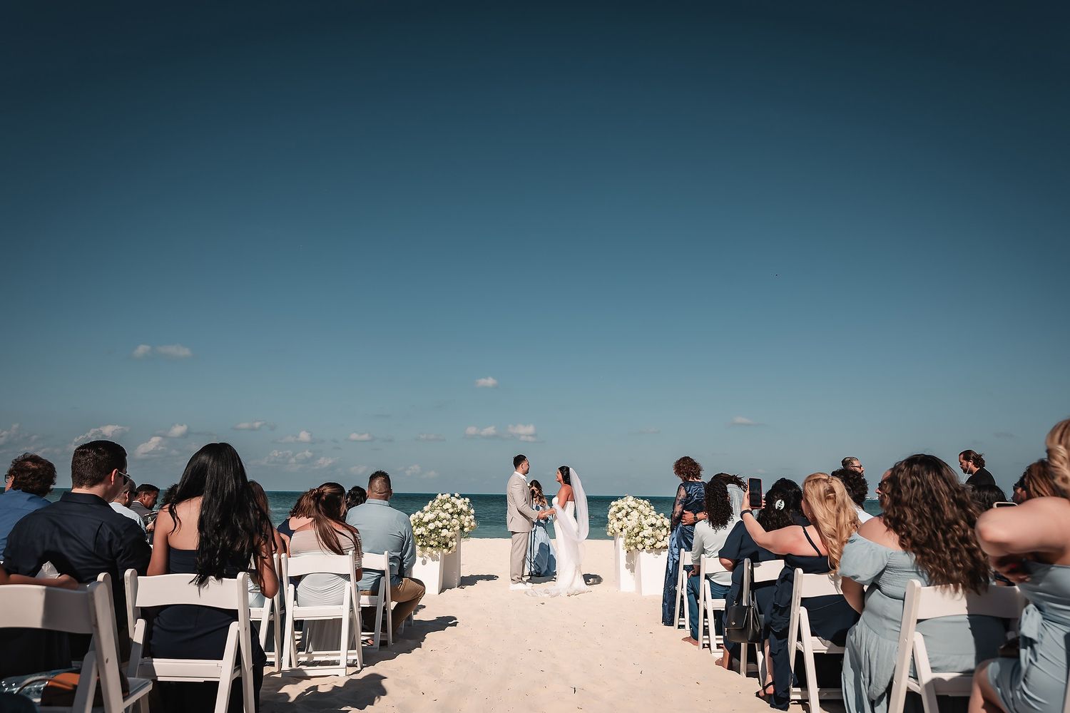 Beach wedding ceremony guests seated in white chairs along the shoreline.
