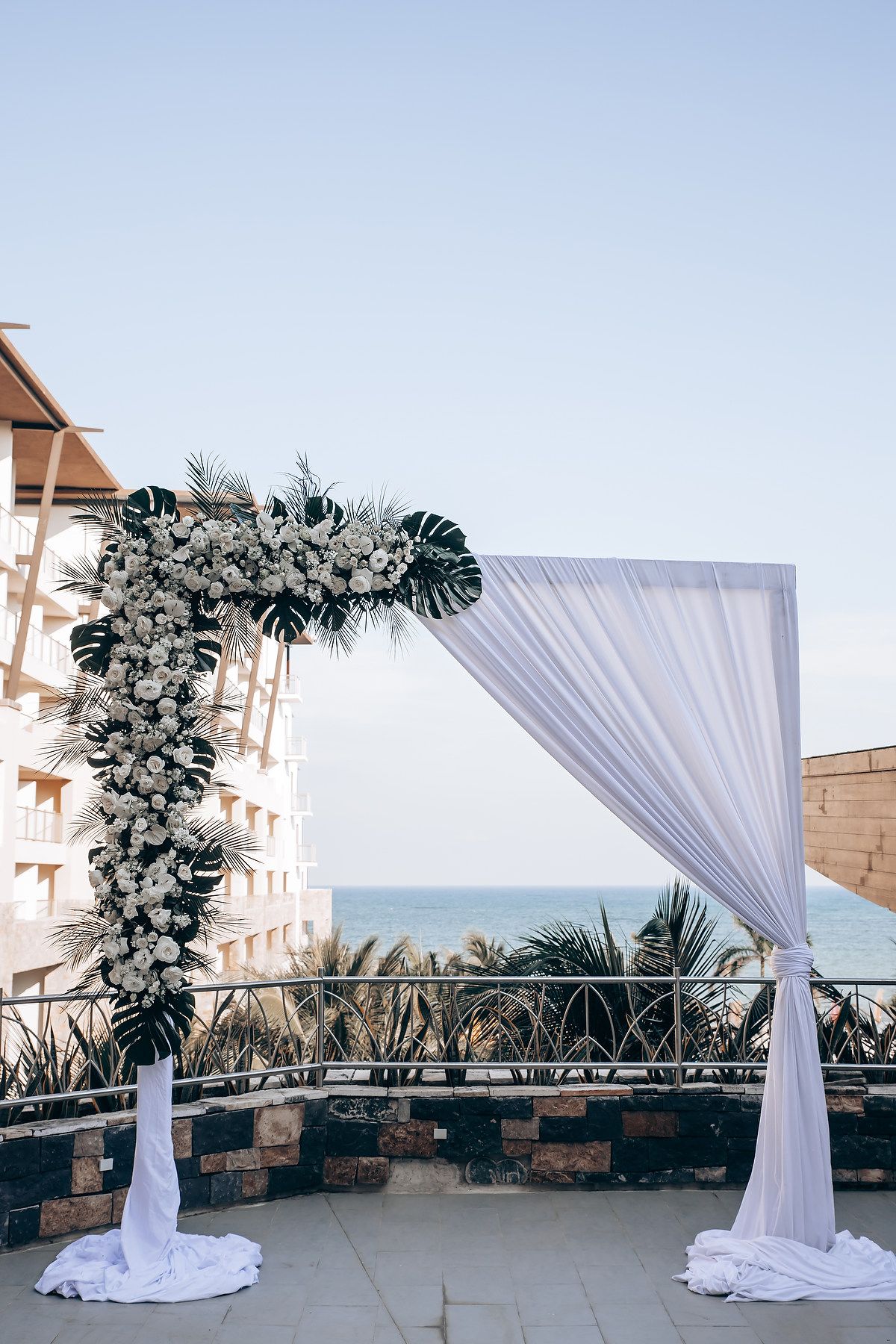 A beachfront wedding arch decorated with white fabric and tropical floral arrangements.