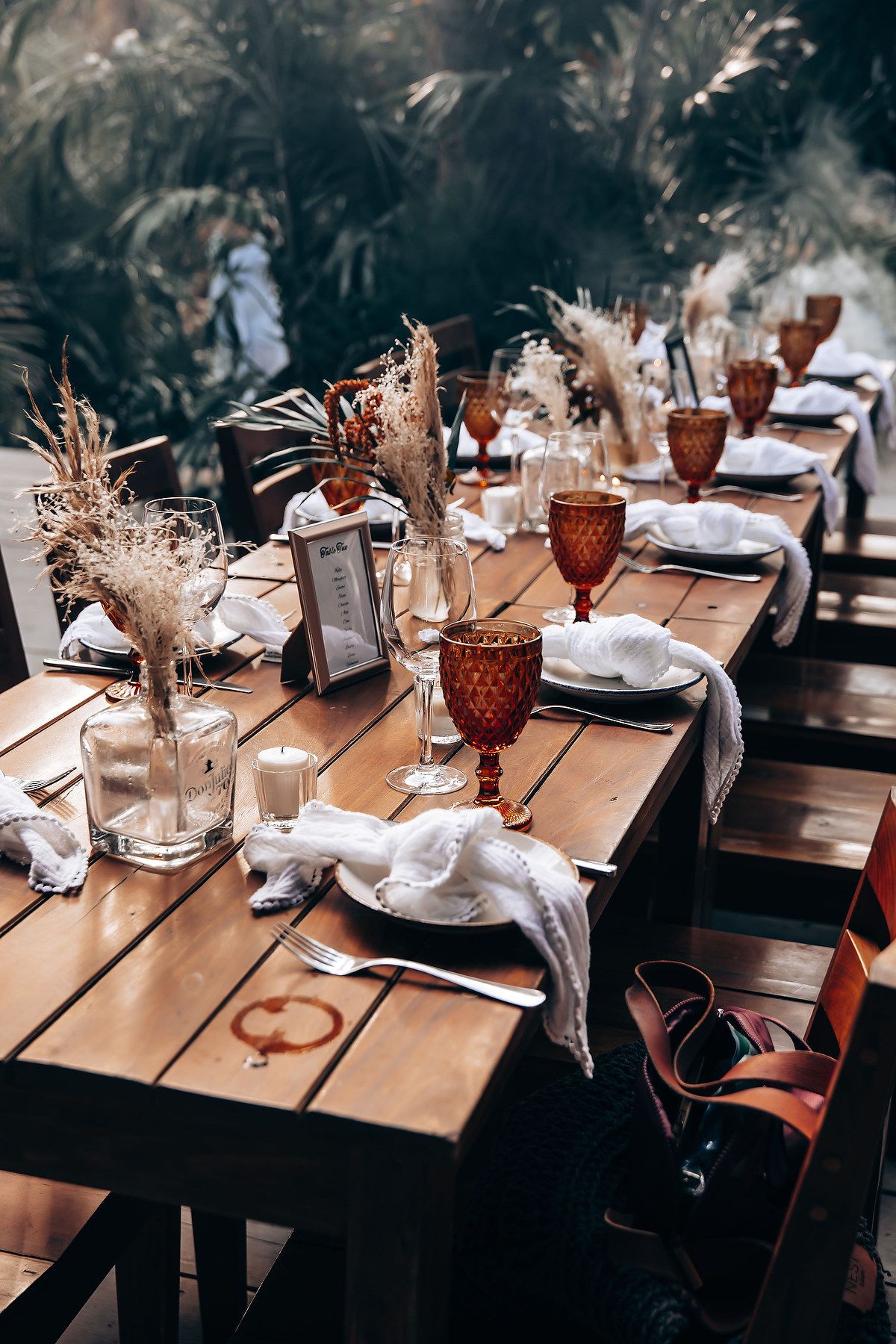Long wooden dining tables set with amber goblets and dried floral centerpieces in a moody restaurant setting.