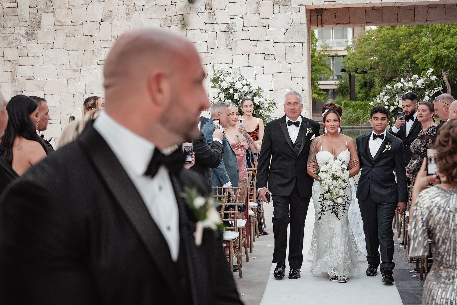 Wedding guests walking in formal attire against a white stone wall backdrop.