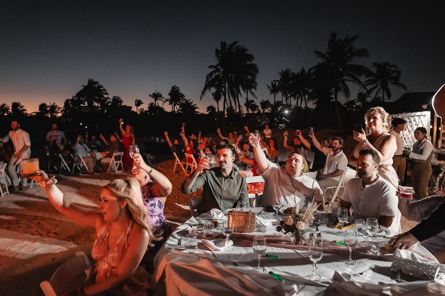 Wedding reception guests celebrate with raised hands during an outdoor evening celebration.