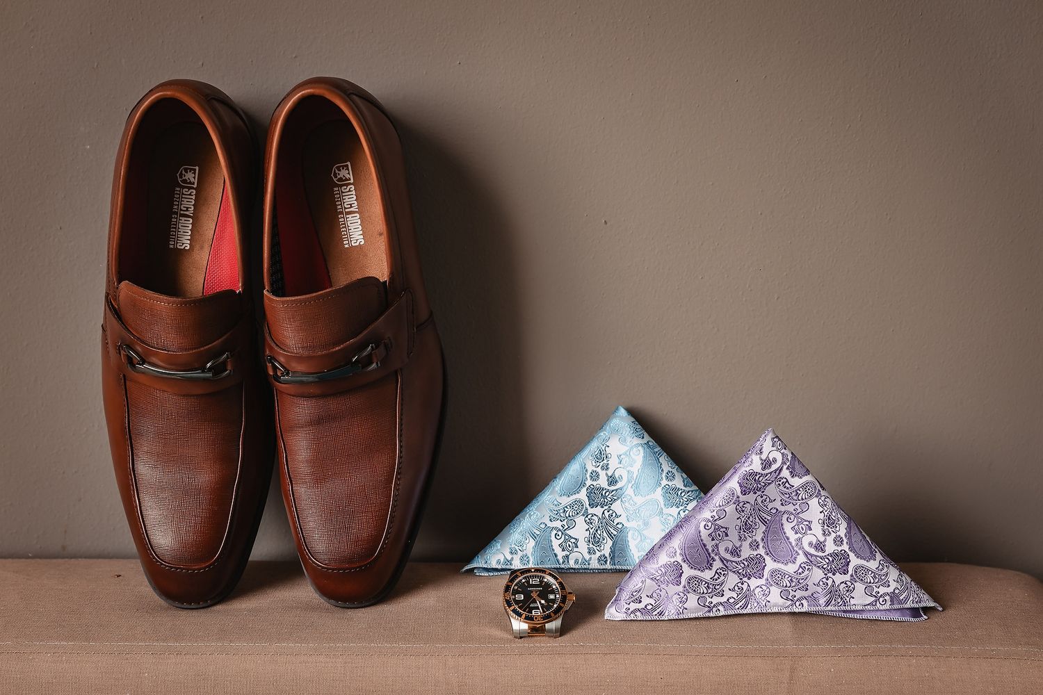 Brown leather loafers displayed beside floral pocket squares and a decorative ring for a wedding.