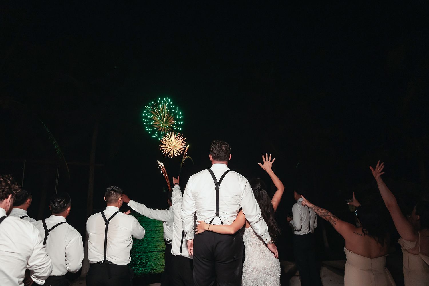 Wedding party watches colorful fireworks light up the night sky during an outdoor beach wedding celebration.