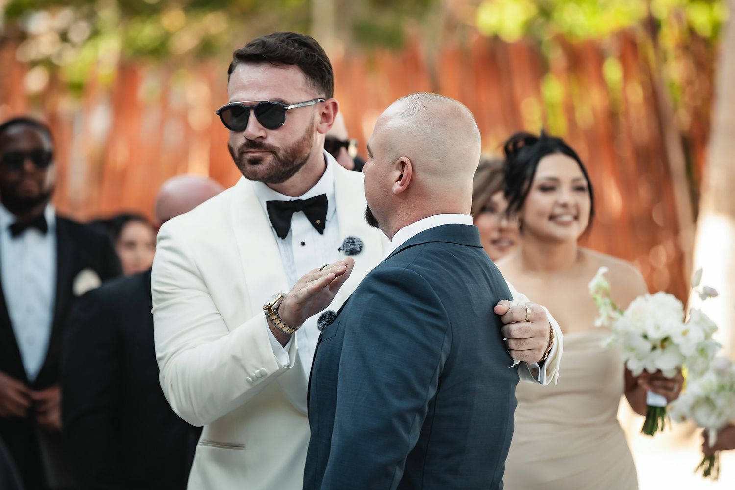 Two grooms share an emotional moment during their outdoor wedding ceremony, one in a white tuxedo with sunglasses.