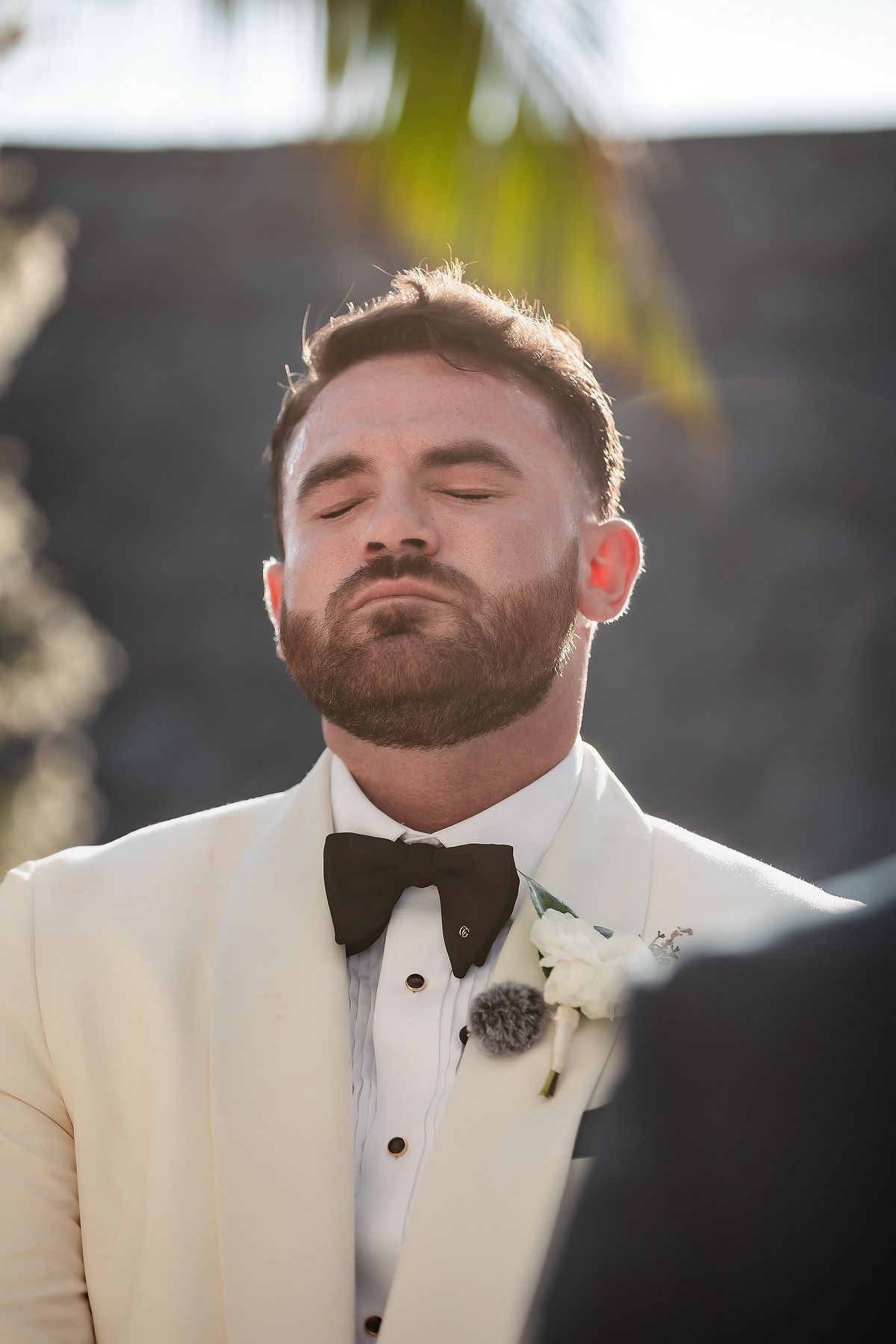 Groom in ivory tuxedo with black bow tie and boutonniere, eyes closed, emotional moment during outdoor wedding ceremony.