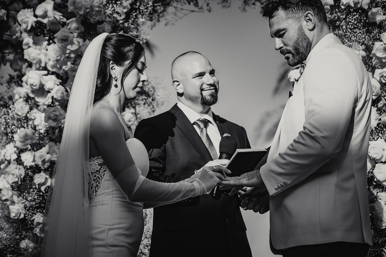 Black and white photo of a couple exchanging vows with an officiant at a floral wedding altar.