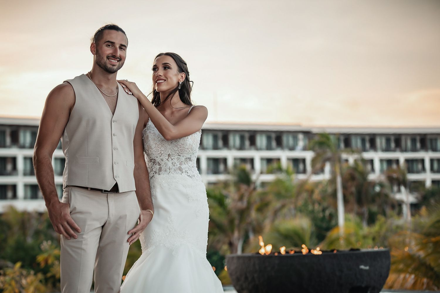 Bride and groom posing outdoors at sunset near a fire bowl with a tropical resort in the background.