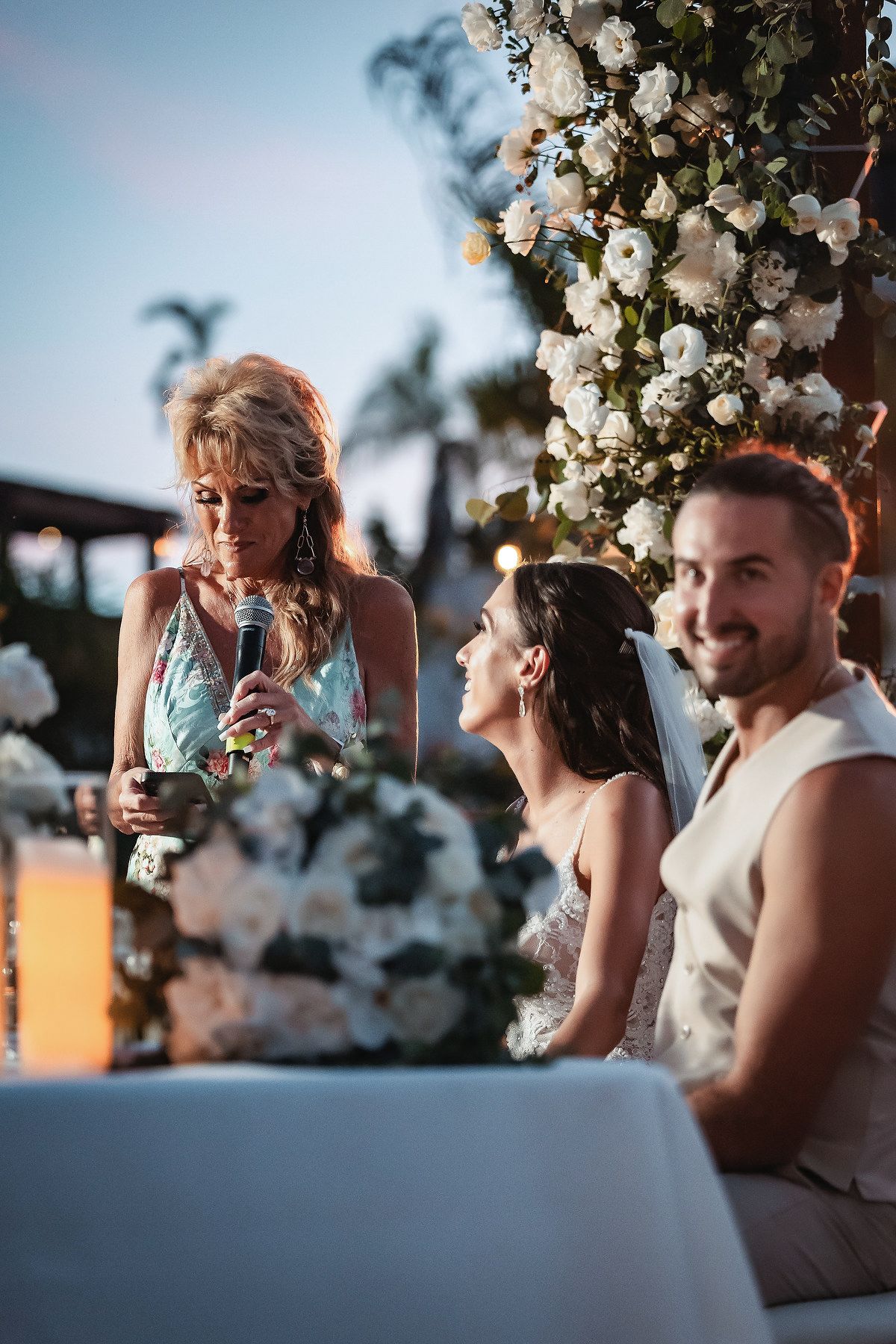Bride and groom smile during outdoor wedding ceremony as woman gives speech near white floral arch at dusk.