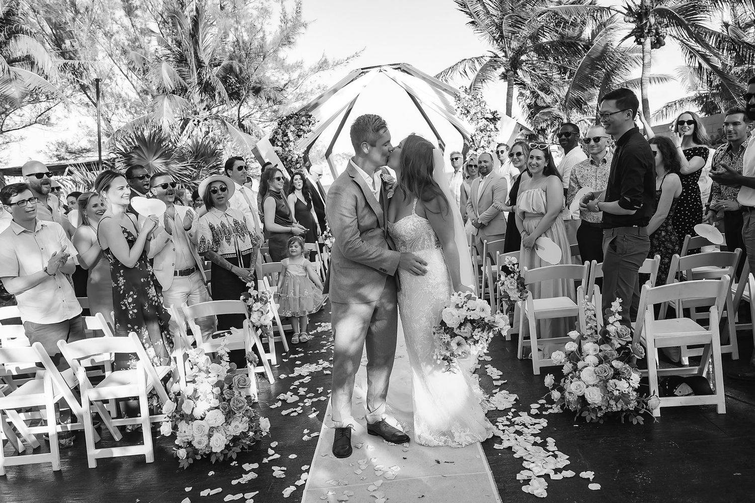Black and white photo of newlyweds sharing a kiss at outdoor wedding ceremony with guests cheering.
