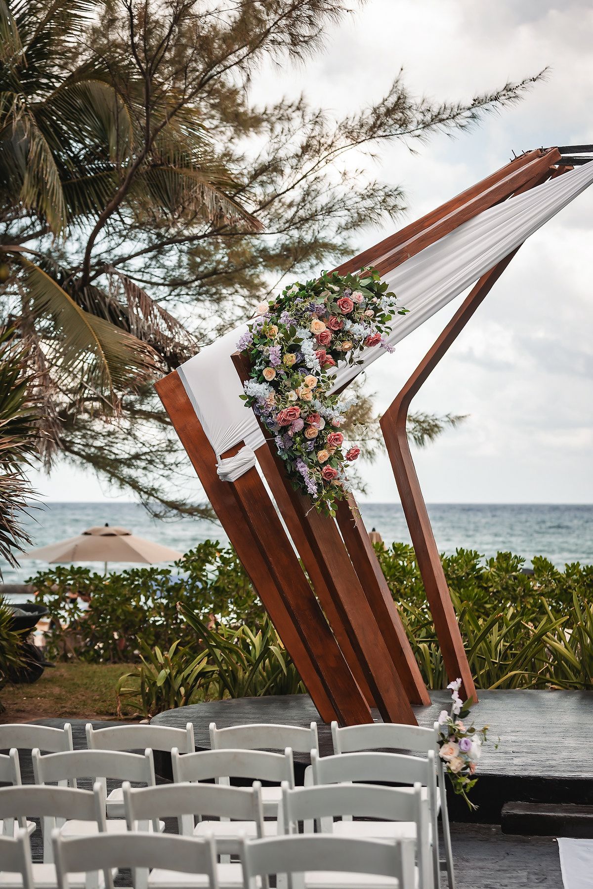 Asymmetric wooden wedding ceremony arch with pastel floral decor and white fabric beside white chairs near tropical beach.