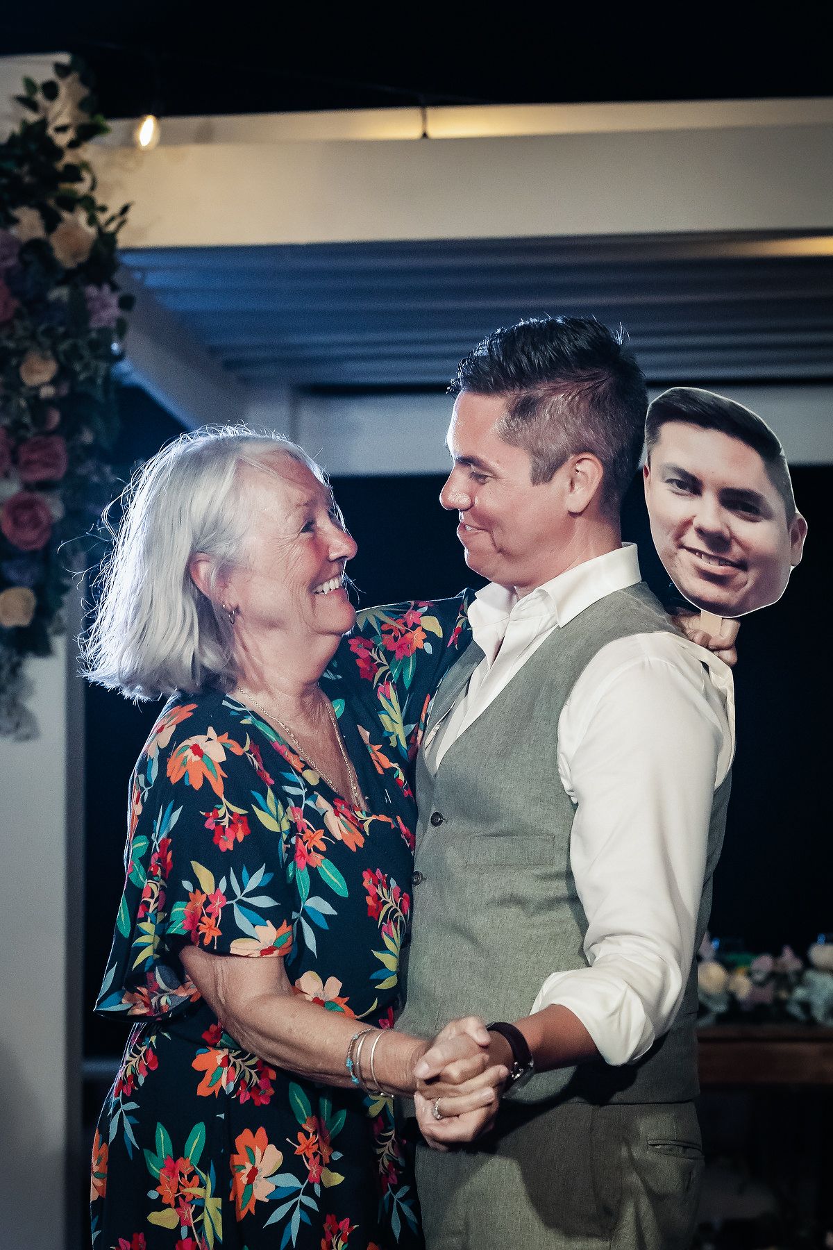 A young man dances with an older woman at a wedding, holding a cutout face prop, surrounded by floral decorations.