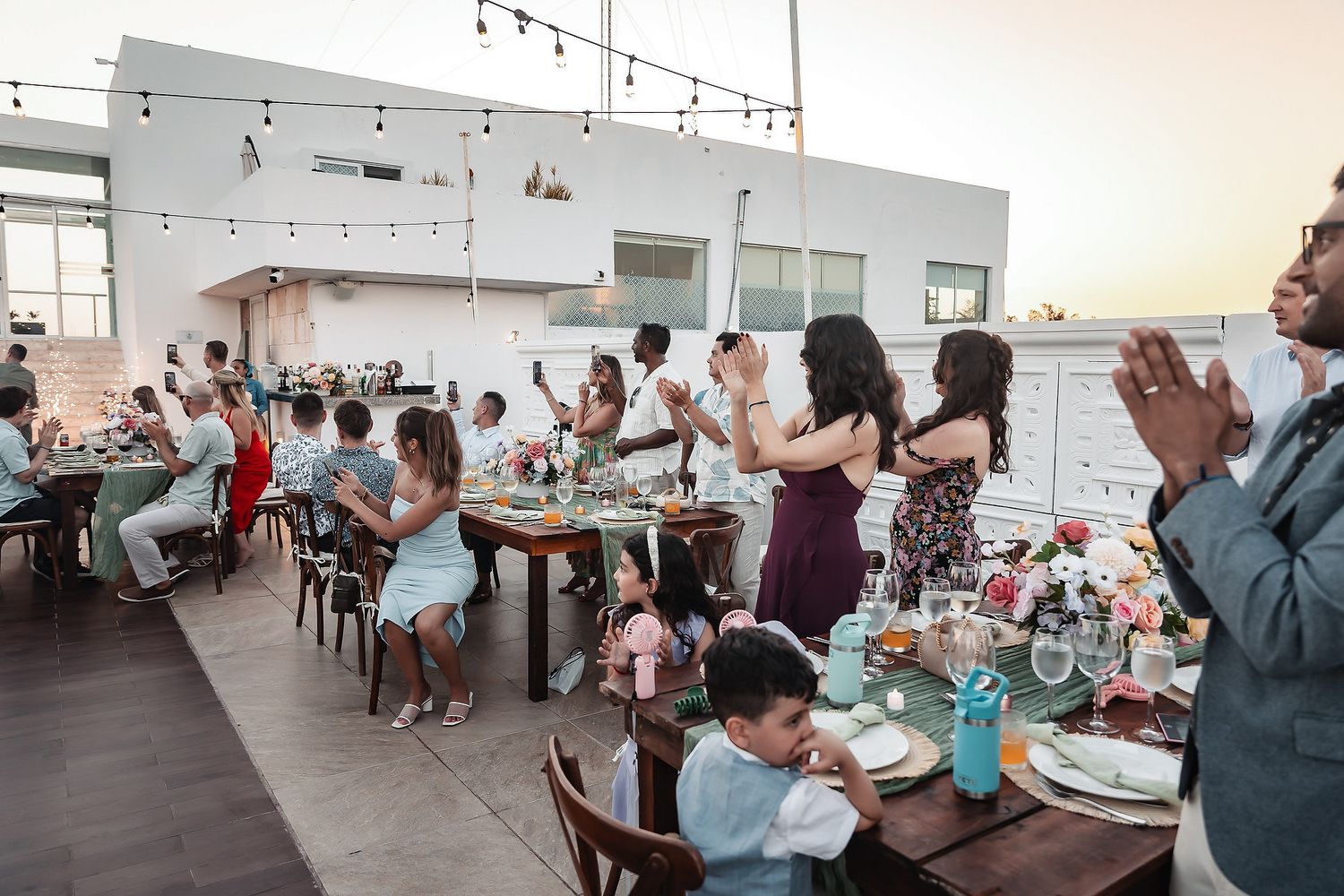 Guests celebrate at an outdoor rooftop dinner party with string lights, floral centerpieces, and a warm sunset backdrop.